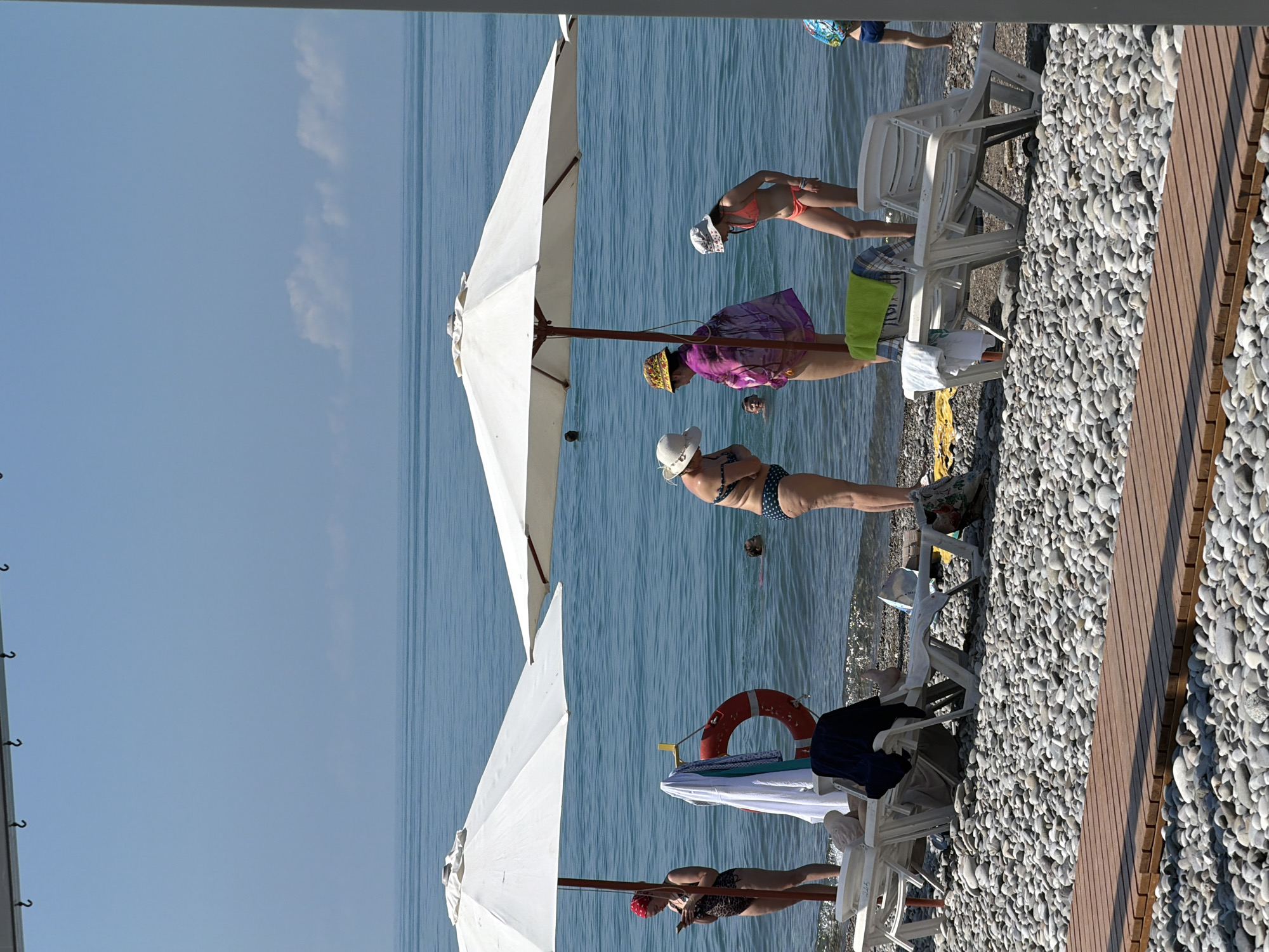 On a sunny day at a pebble beach in Sochi, Russia, several people are enjoying the Black Sea. Two large white umbrellas provide shade for beach chairs. A woman in a leopard-print swimsuit stands near the water, looking at her phone. Another woman in a polka-dot bikini stands on the shore. A third woman in a patterned wrap and a hat converses with a woman in an orange bikini, who is looking down. A man is visible in the water. A life preserver hangs from a pole. The sea is calm and blue, with a few fluffy clouds in the distance. A wooden walkway extends across the foreground.