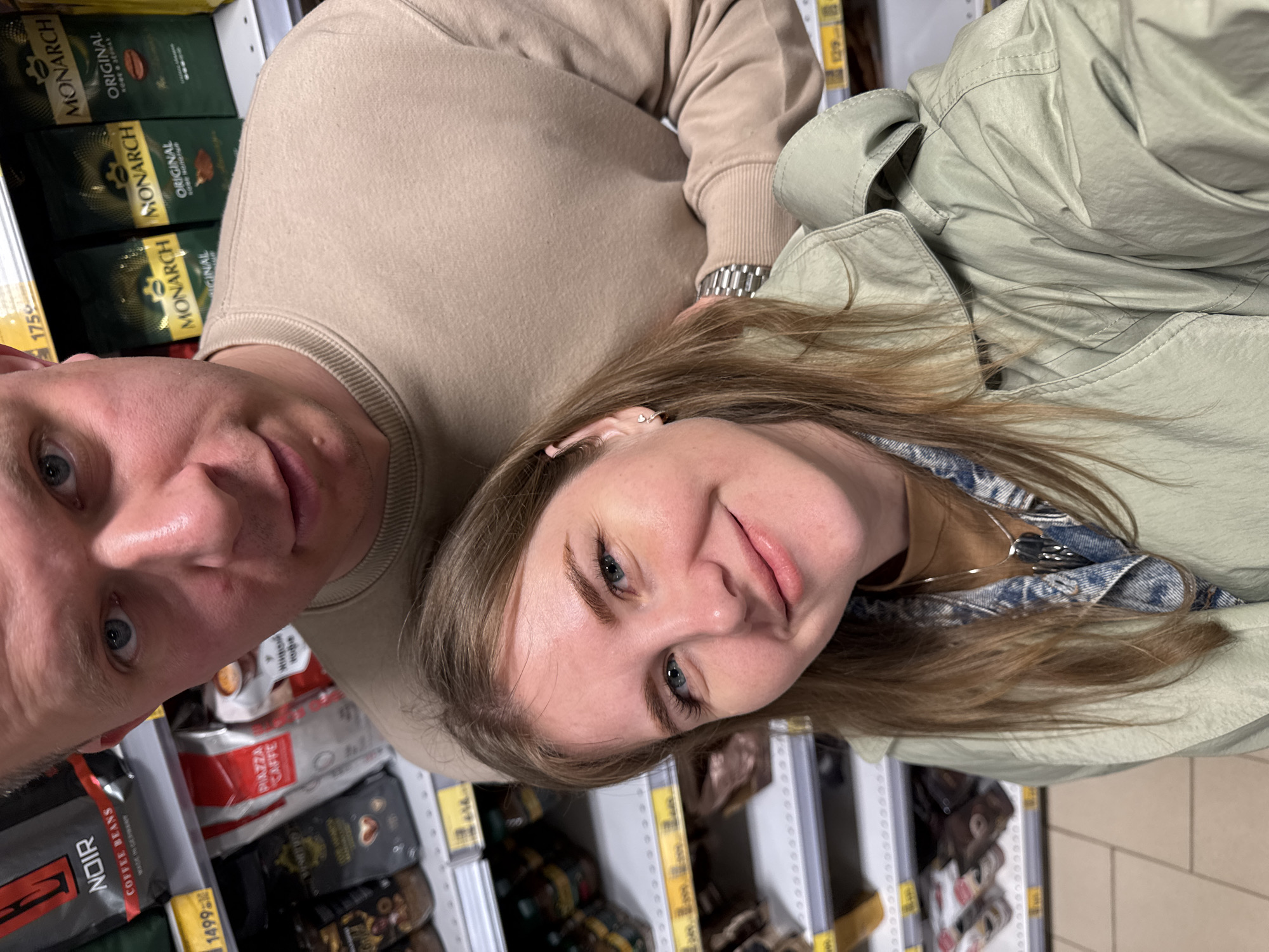 Here is a detailed description of the image:
**Scene:** The image appears to be a selfie taken by a couple inside a grocery store. They are standing in an aisle with shelves stocked with coffee products.
**People:** A man and a woman are present. The man is closest to the camera, showing the left side of his face with blue eyes. The woman is next to him with her face clearly visible. She also has blue eyes and is smiling slightly.
**Subjects:** The main subject is the couple taking the selfie. Additionally, coffee products on the shelves are also significant.
**Objects:** Key objects in the scene include bags and cans of coffee (brands like "Monarch", "Caffe"), clothes (such as a sweater and a jacket), and the metal shelves with price tags.
**Actions and Interactions:** The primary action is the couple taking a selfie. There is likely an interaction happening between them (e.g., selecting items in the store).
**Environment:** The scene is set inside a grocery store, with well-lit aisles and shelves full of coffee products. The image is rotated, so vertical lines appear slightly angled.
**Location:** There is no indication in the picture that the location is Горный, Russia. If location information is needed, that will require data from outside the image itself.