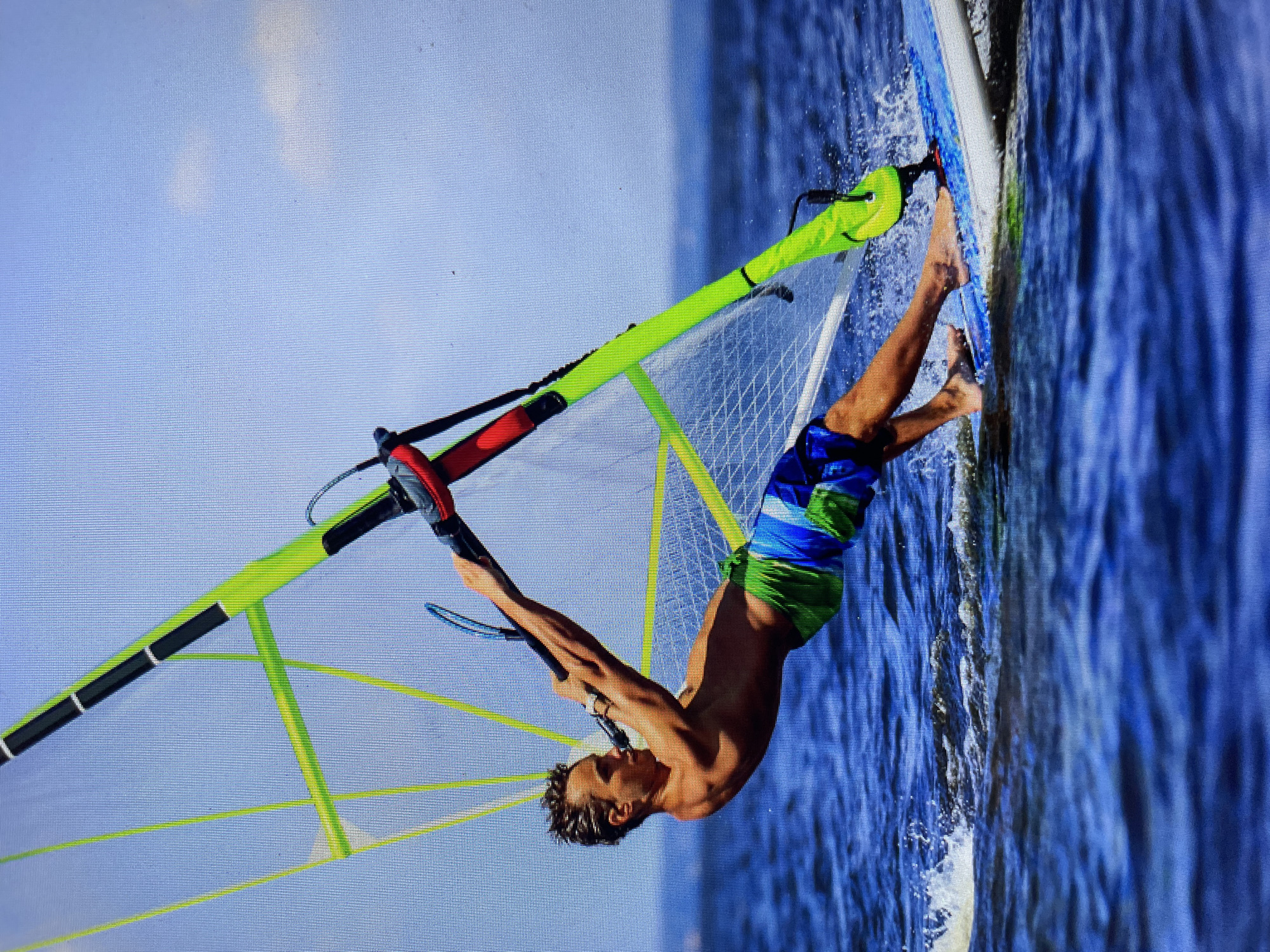 A male windsurfer is depicted in action on the water, presumably in the city of Bacong, Philippines. The scene shows the shirtless man wearing blue and green shorts, gripping the windsurf sail while positioned on the board. His body is angled downwards toward the water. The sail is vividly colored, with a green frame and a mesh interior. The windsurfing board is white. The background features the blue water of the sea or ocean under a bright sky.