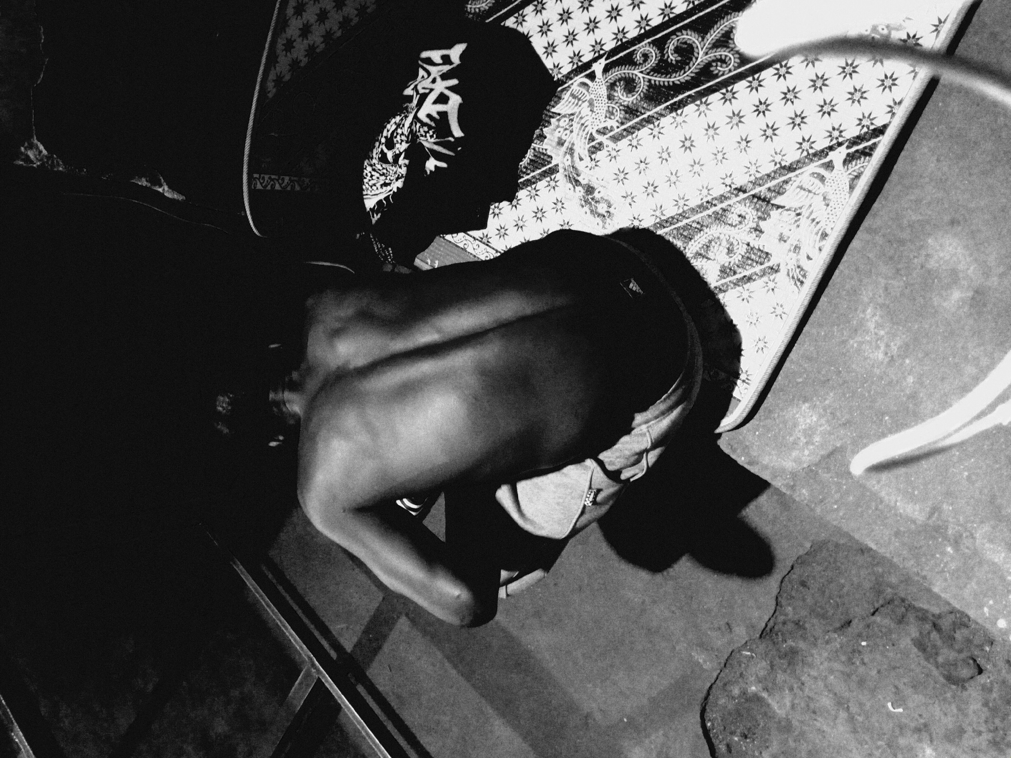 A high-angle, black and white photograph depicts a shirtless person in the foreground, leaning over at the waist. They are wearing jeans, and their face is obscured. To the right is a patterned mat or blanket. There is a dark object with designs on top of the blanket. Parts of a metal structure appear in the bottom left of the frame, and what looks like a rope or cord is in the top right. The ground has a rough texture.