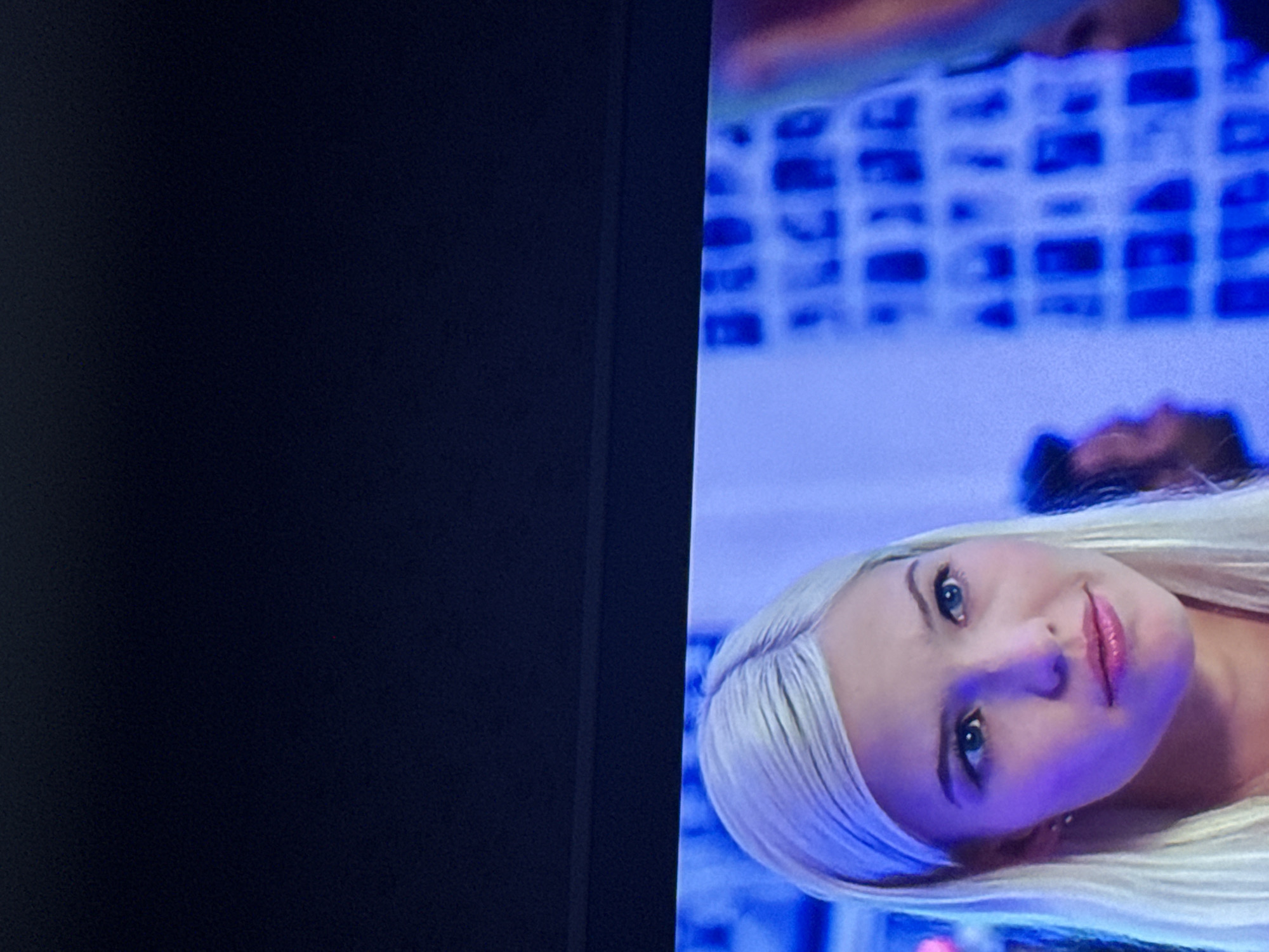 The image shows a television screen displaying a woman with blonde hair and blue eyes, looking towards the camera. Behind her is a blurry background with a blue hue and what appears to be a grid-like pattern. Part of the black television frame is visible on the left side of the image.
