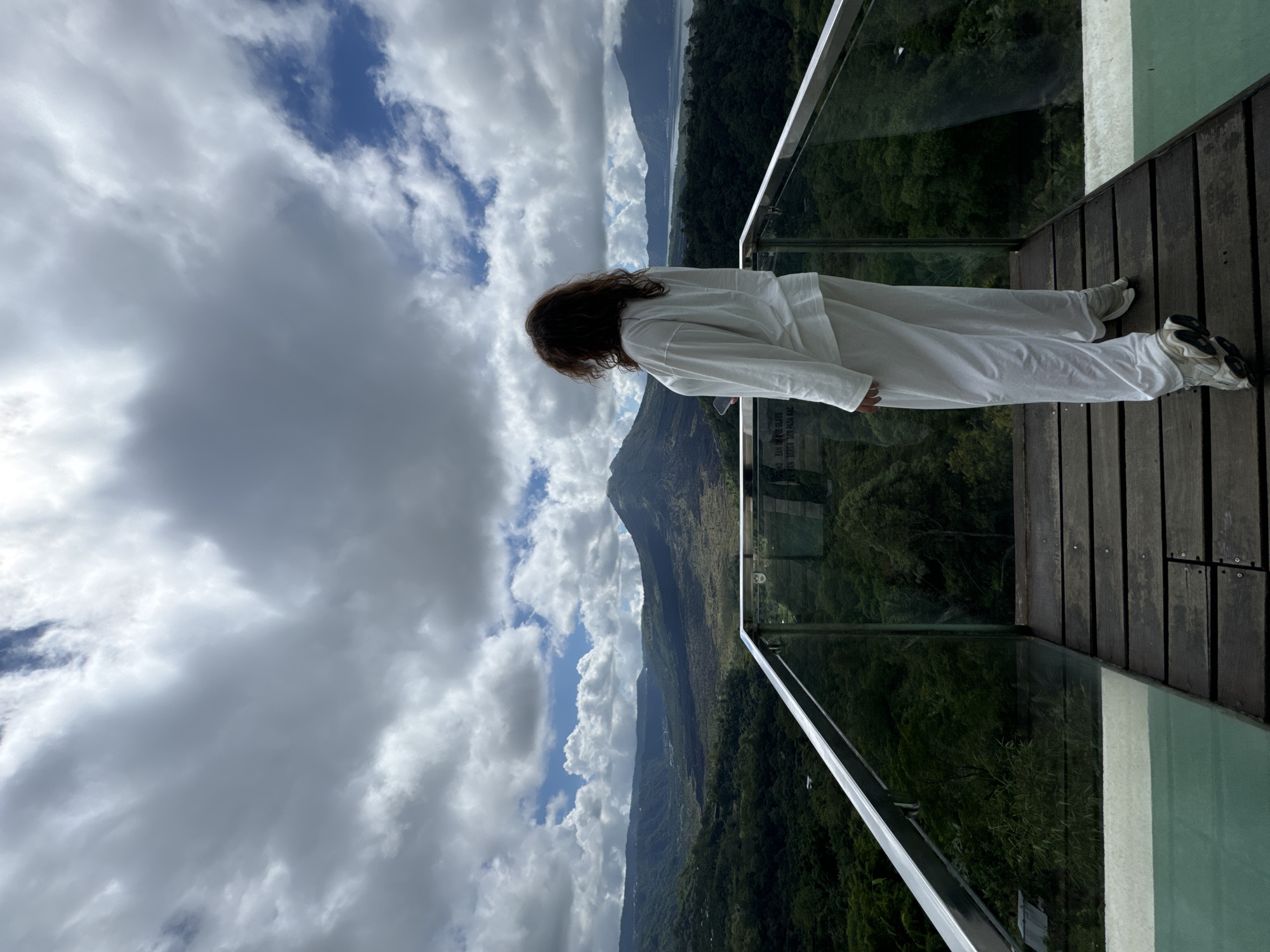 A person is standing on a wooden deck overlooking a mountainous landscape in Kintamani, Indonesia. The person is wearing white clothing and faces away from the camera, looking out at the view. The deck has a glass barrier. The sky is cloudy with patches of blue. Mountains covered in green vegetation stretch into the distance.