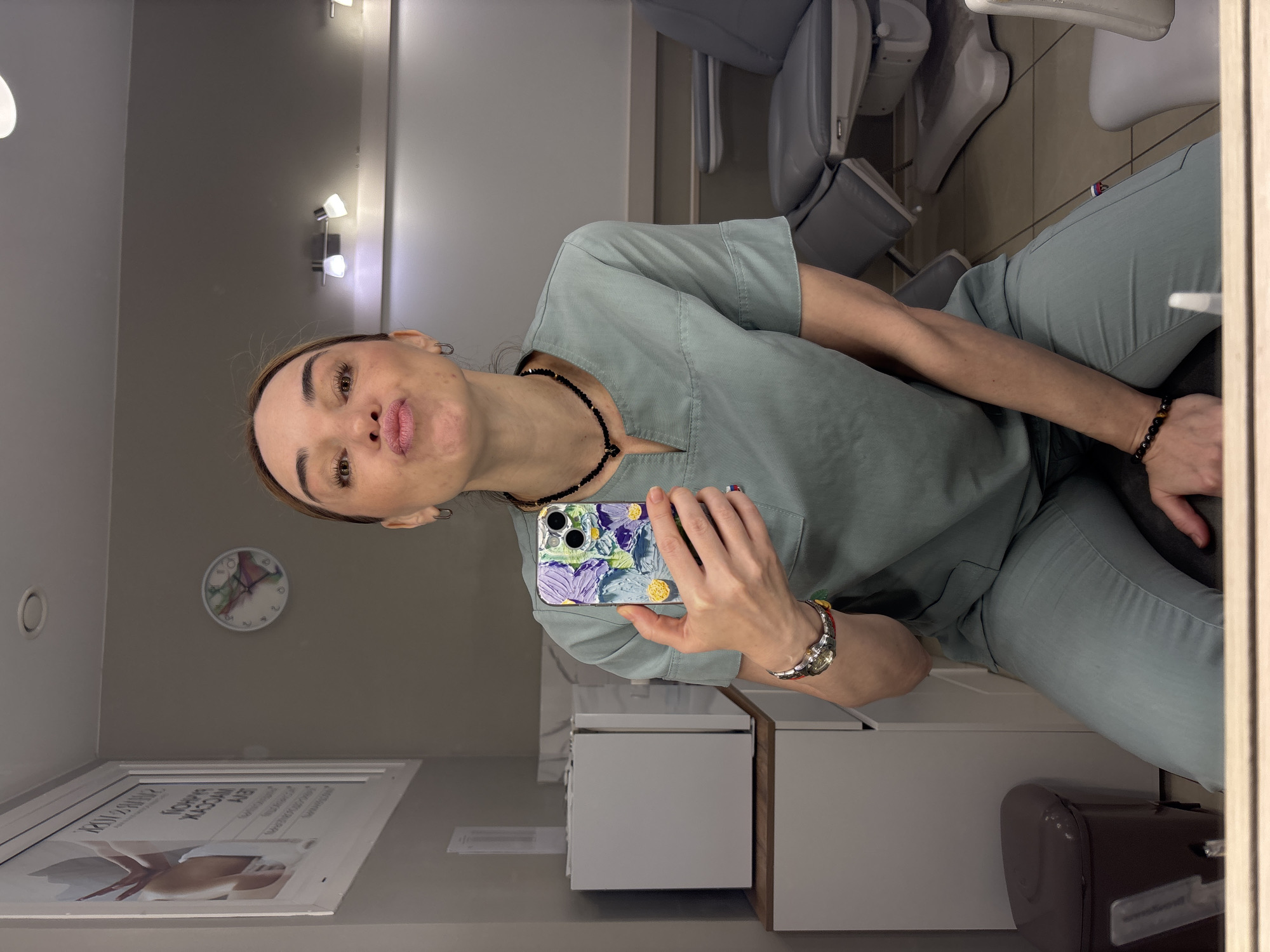 The image captures a mirror selfie of a woman in what appears to be a professional setting, possibly a clinic or spa. The woman, who has light brown hair and dark eyes, is wearing light green scrubs and is making a "fish face" expression by puckering her lips. She holds a smartphone with a distinctive textured floral case, featuring purple, green, and yellow elements. She also wears a black beaded necklace, a matching black beaded bracelet on her right wrist, and a silver watch with a colorful band on her left wrist.
The background shows a brightly lit, modern room. To her left, a light grey wall features a round clock with an abstract design and a white-framed poster. The poster displays text in Cyrillic, with visible phrases such as "ПАНАМСКАЯ" (Panamanian) and "ПОПНАЯ КОРРЕКЦИЯ ЛЕГ." (suggesting a cosmetic body treatment like "butt correction"). A circular vent is also visible on this wall. To her right, several grey, ergonomic chairs or treatment beds are arranged. Below her arm, a white cabinet or storage unit is visible, and a dark bin is partially seen at the bottom right. Multiple wall-mounted lights illuminate the space. The scene indicates an indoor environment, likely a professional wellness or beauty establishment.