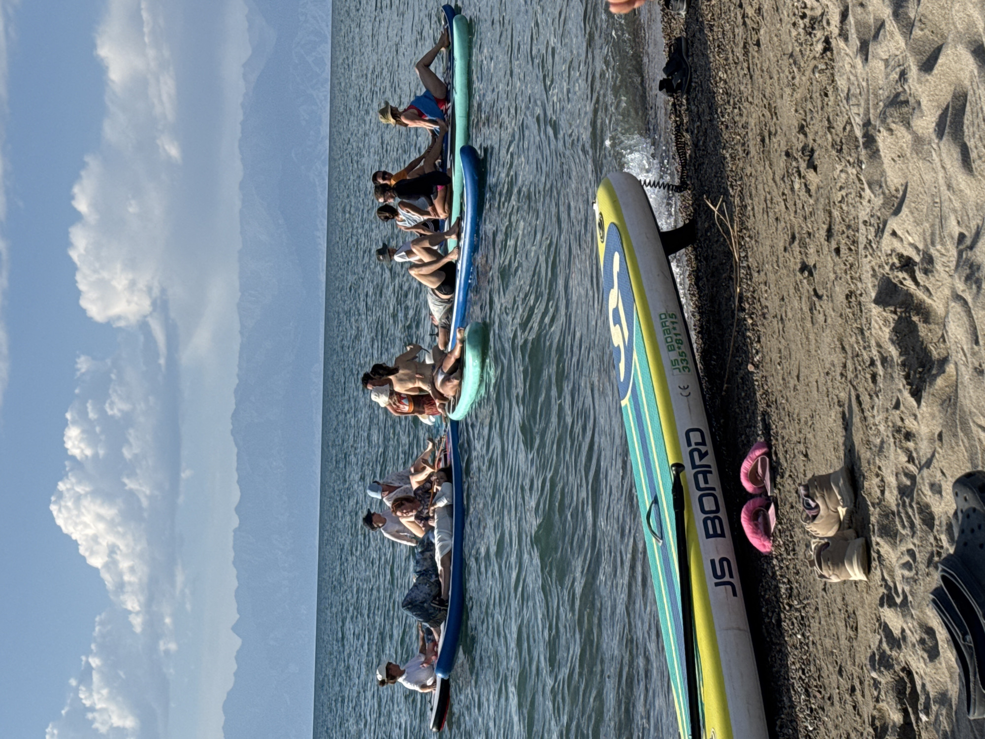 A group of ten individuals is seen scattered across multiple paddleboards on a body of water, likely Lake Issyk-Kul, near the city of Balykchy, Kyrgyzstan. The individuals are in various states of relaxation and interaction, with some lying down and others sitting or conversing. The water is calm with small ripples, and the sky is partly cloudy with a clear blue expanse. In the background, a mountain range is visible, suggesting a scenic, natural environment. On the shore in the foreground, there are two paddleboards, one of which has the text "JS BOARD" printed on it, along with a paddle and some personal items like shoes and flip-flops. The overall scene depicts a leisure activity in an outdoor setting.