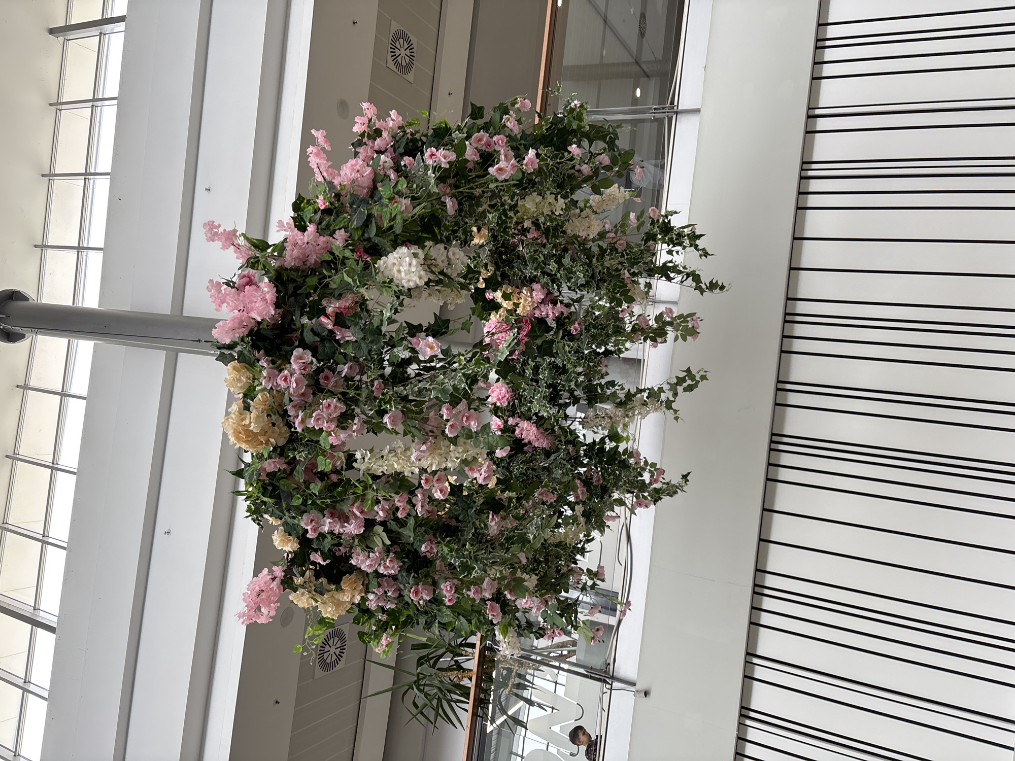 A low-angle shot shows a floral arrangement suspended from a gray support pole within a building, likely indoors. The arrangement features pink, white, and pale yellow flowers intermixed with green foliage. The background includes white walls with black horizontal stripes on one side and a window with a grid pattern and white vertical panels on the other. A ventilation grille is visible near the flowers. Below the flowers, vague figures can be seen through the window. The photograph does not offer explicit information to directly confirm its exact location in Burgas, Bulgaria.