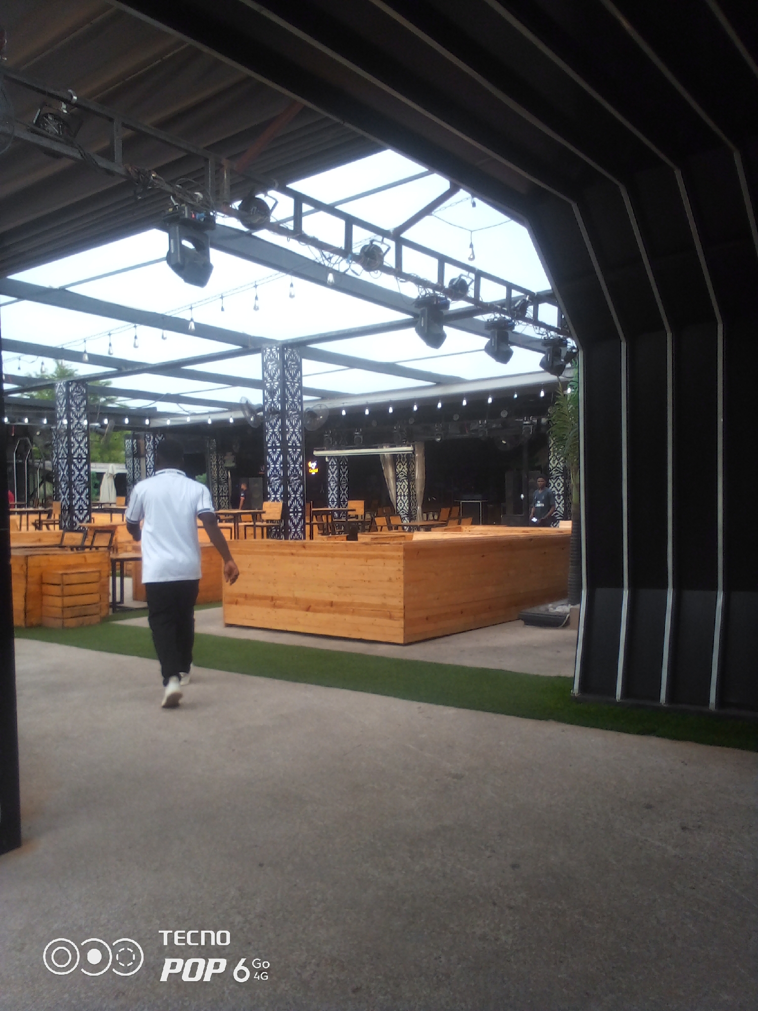 The image depicts an outdoor seating area at night in Howayi, Nigeria. The scene features a covered patio with a metallic roof structure supported by pillars. Several spotlights are suspended from the roof. The area is furnished with wooden benches and tables, with some areas covered by artificial turf. A man in a white shirt and black pants is walking away from the camera. Other patrons are visible in the background, some seated at tables. The overall ambiance suggests a venue for dining or entertainment. The TECNO POP 6 GO 4G watermark is present in the lower right corner.