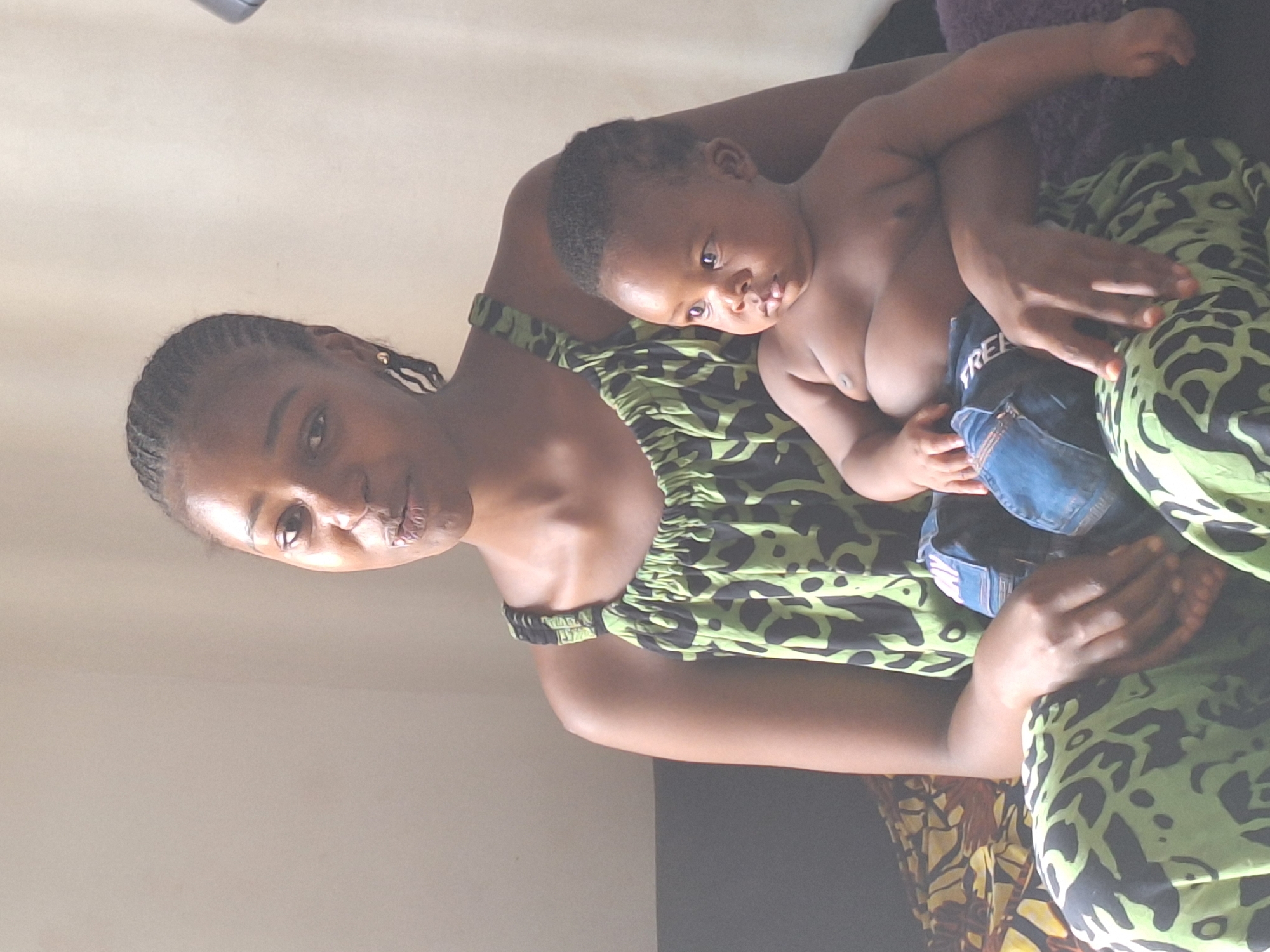 The image, taken in Sabon Tasha, Nigeria, depicts a mother holding her baby in an indoor, intimate setting, likely a home. The woman, with dark skin and braided hair styled in cornrows, is positioned on the left side of the frame, looking directly at the viewer with a neutral to slightly thoughtful expression. She wears a lime green and black patterned strapless or off-the-shoulder dress or top and has a small gold earring visible.
She cradles a baby, also with dark skin, who is shirtless and appears to be wearing blue denim shorts or a diaper cover. The word "FREE" is partially visible on the baby's denim clothing. The baby looks towards the camera with a calm and curious gaze. The scene suggests daytime, with soft, even lighting, though specific time-of-day cues like windows are not visible.
In the background, a plain, light-colored wall occupies the upper and left portions of the image. Towards the lower right, darker fabrics or furniture are visible, including a hint of a purple textured cloth and a yellow patterned fabric, suggesting a comfortable, domestic environment. The overall mood is tender and familial.