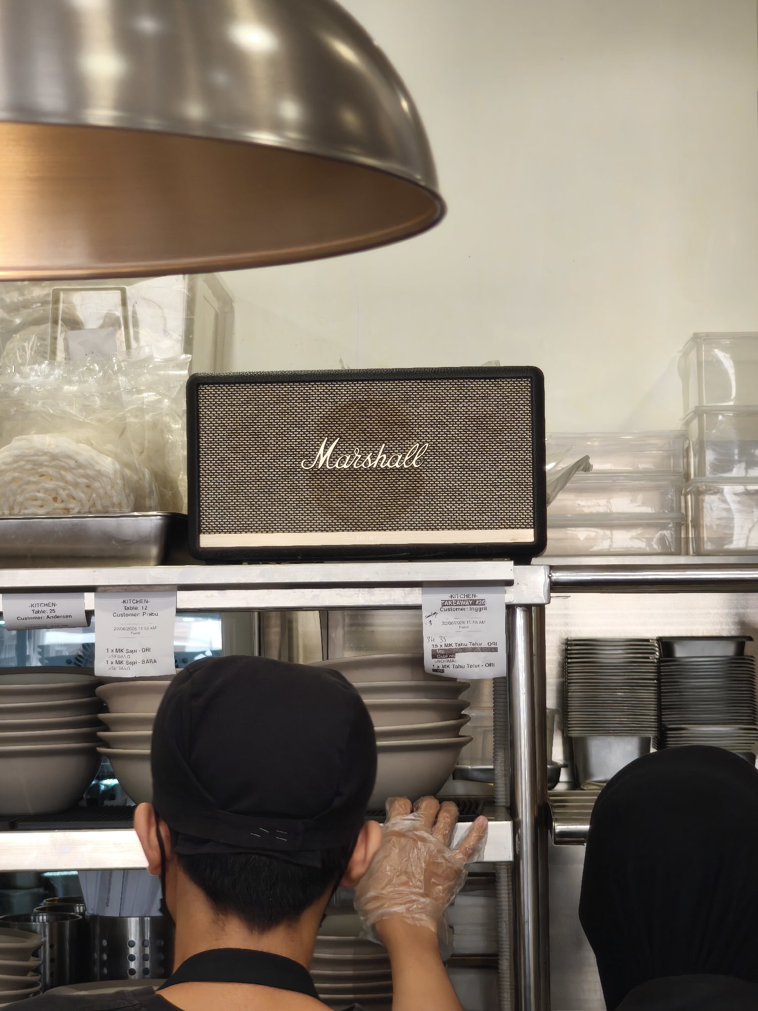 In South Jakarta, Indonesia, a close-up, eye-level shot captures the interior of a kitchen environment. Dominating the center is a black Marshall speaker with a beige speaker grille, positioned on a stainless steel shelf. To the left, stacks of white shredded ingredients are contained in a metal tray, partially obscured by plastic packaging. Below this, white bowls are neatly stacked on another shelf.
In the foreground, the backs of two individuals are visible. The person on the left, wearing a black skullcap and apron, has their head turned to the side, revealing their ear. Their right hand, clad in a transparent plastic glove, is resting on the shelf. To the right, the back of another person wearing a black head covering is visible.
Attached to the shelf below the Marshall speaker are two white order slips. The slip on the left reads "KITCHEN," "Table: 2," "Customer: Andersen," and "22/06/2023 11:53 AM." The slip on the right also has "KITCHEN" and "TAKEAWAY #26," "Customer: Ibrahim," and "22/06/2023 11:53 AM." Below this, it lists order details such as "15 X MK Tahu Telur - ORI," "1 X MK Supi - BARA," and "1 X MK Tahu Telur - ORI."
In the background, additional shelves hold stacks of clear plastic containers and black containers, suggesting a busy food preparation or service area. A large, reflective metal lamp fixture hangs from the ceiling, casting a warm light on the scene.