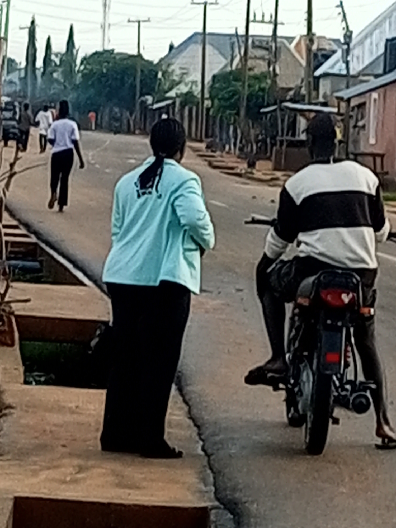 The image depicts a street scene in Tumfure, Nigeria, characterized by its residential and commercial architecture. In the foreground, a person is walking on a paved sidewalk while another person is seated on a motorcycle. The person walking is wearing a light blue jacket with a black design on the back and black pants. The person on the motorcycle is wearing a black and white striped long-sleeved shirt and dark pants. The motorcycle is black and is positioned on the asphalt road. In the background, several other individuals are visible walking along the road, and a few structures, including buildings and stalls, line the street. The overall environment suggests a typical day in a Nigerian town, with everyday activities taking place on the street.