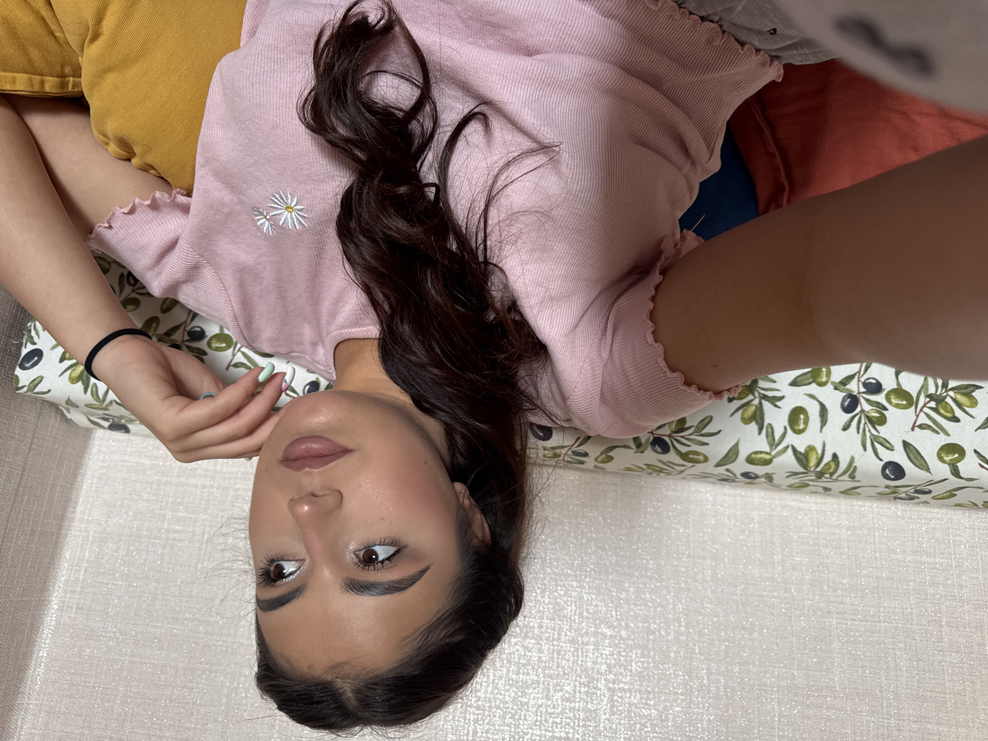 A young woman is lying down indoors on patterned cushions. She is wearing a pink, textured shirt with a daisy design and has long, dark hair. She has fair skin and appears to be looking upwards. The cushions beneath her head are patterned with olives and olive branches. There are additional pillows behind her, including yellow and coral ones. The location is unknown and cannot be determined from the visual data.