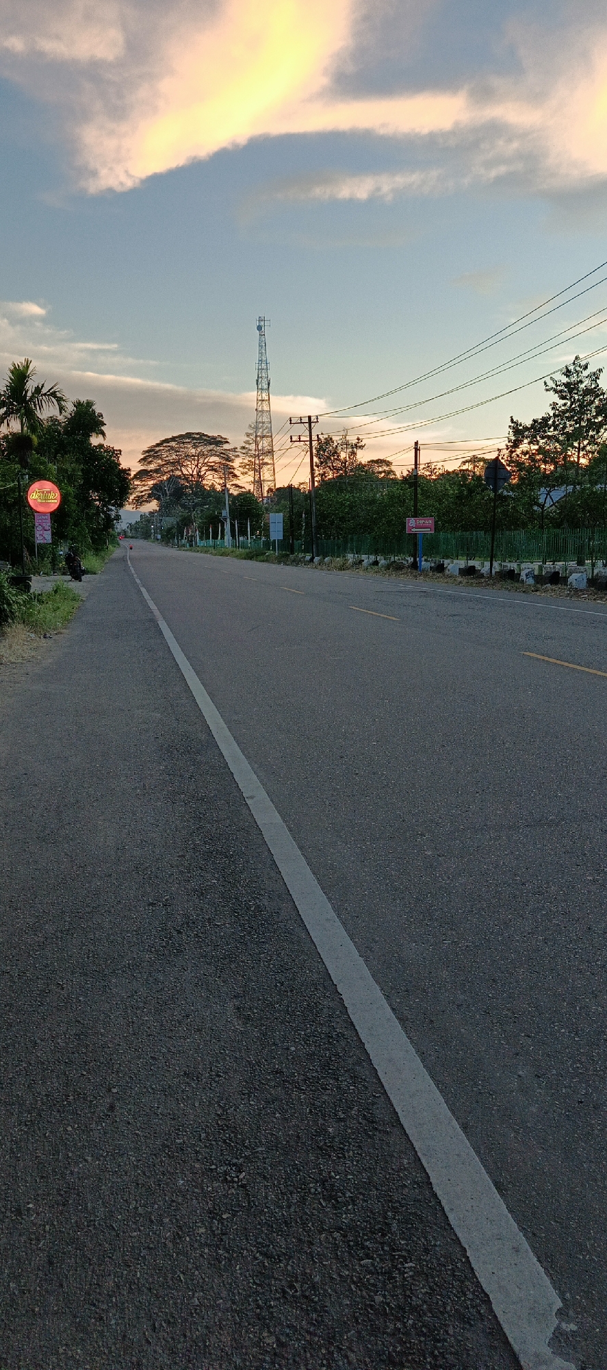 The image shows a paved road in Sukamulia, Indonesia, during what appears to be sunset or sunrise. The sky is a gradient of blue with bright yellow and pink clouds. On the left side of the road, there are trees, a palm tree, and a circular red sign with white text that reads "Deruik". Further down the road, a tall telecommunications tower stands against the sky, accompanied by utility poles and wires. The road has a white dashed line marking the lane and a yellow line on the right side. The environment is rural or semi-rural, with lush green foliage lining the sides of the road. The perspective is from a low angle, looking down the road. No people or vehicles are visible in the frame.