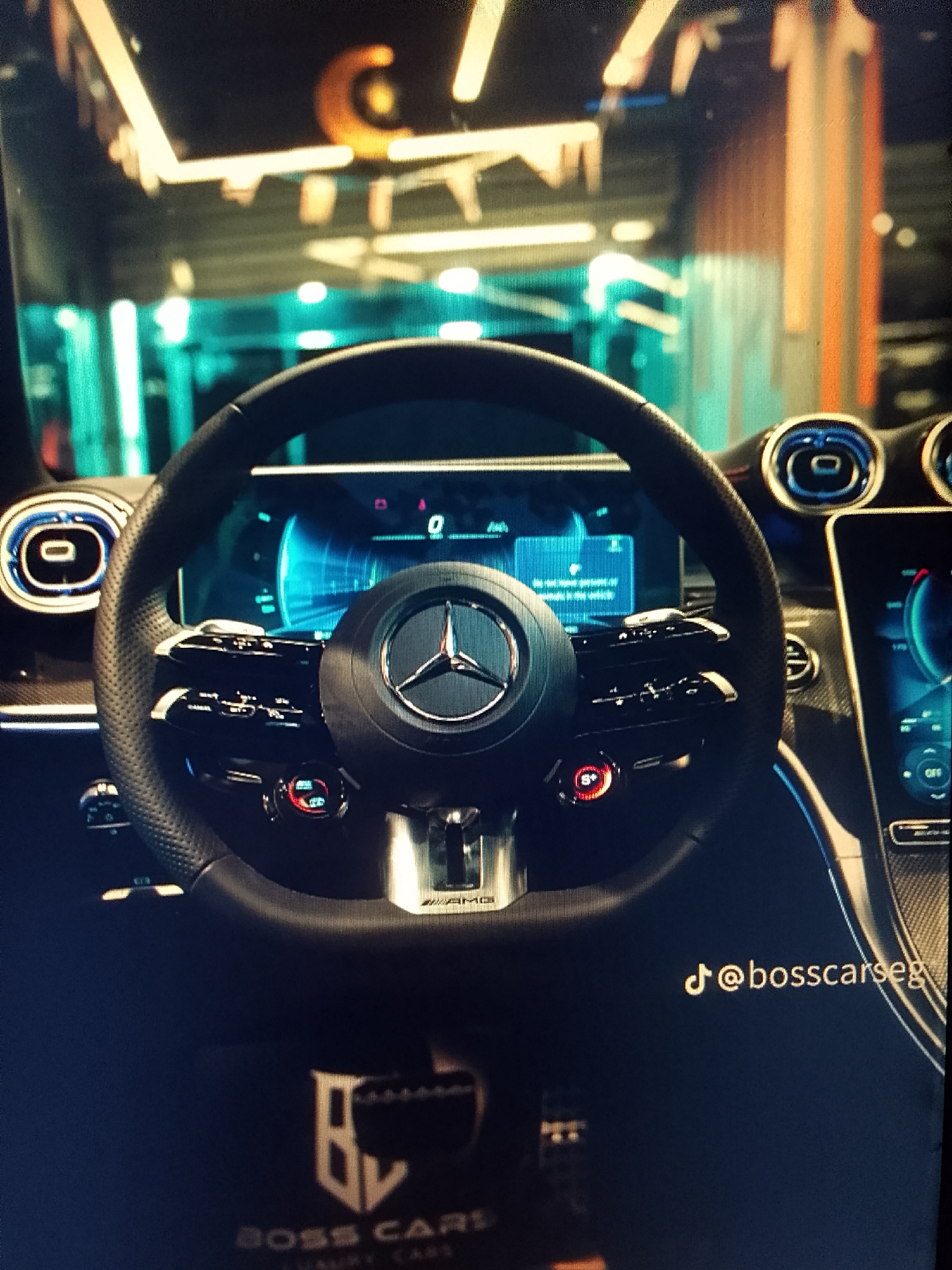 The image captures the interior of a stationary Mercedes-Benz AMG vehicle, focusing on the steering wheel and dashboard from the driver's perspective. The multi-function steering wheel, crafted with perforated dark material, features a prominent silver Mercedes-Benz star emblem in the center and "AMG" branding at the bottom spoke. Two circular control dials are affixed to the lower part of the steering wheel: one is bright red and displays "S+" (Sport Plus), while the other is dark with a silver rim. The digital instrument cluster behind the steering wheel is illuminated with blue graphics, displaying a speed of "0" and a partially visible warning message: "Do not leave persons or...". To the right, a large vertical infotainment screen shows a blue and red graphical interface, with "OFF" visible on one control. Circular air vents with illuminated blue rings flank the instrument cluster. The dashboard appears to incorporate dark, possibly carbon fiber, trim. The immediate foreground below the steering wheel displays a faint logo: "BV BOSS CARS LUXURY CARS". In the bottom right corner, a TikTok handle, "@bosscarseg", is visible. The background, seen through the windshield, is blurred but suggests an indoor environment, possibly a showroom or garage, with various illuminated structures, vertical light fixtures in turquoise and orange hues, and a large orange "C" or similar graphic element on a wall or ceiling. The location is Unknown, Unknown.