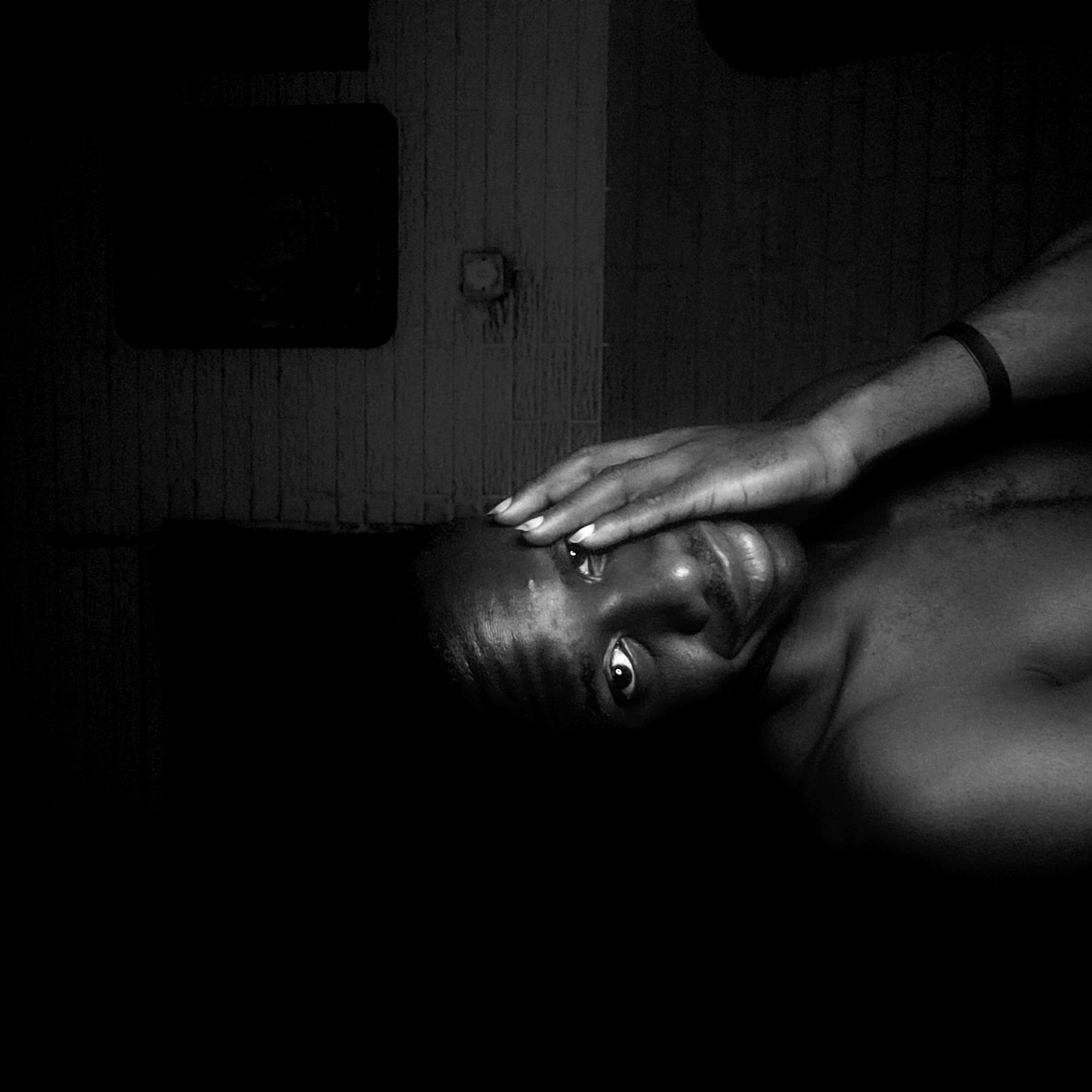 A close-up, black and white photograph shows a shirtless man lying on his side. His right arm is raised, with his hand resting on his forehead. He appears to be in a bathroom in Narayi, Nigeria, with tiled walls visible in the background, including a dark square object and an electrical fixture. The lighting is low, with deep shadows obscuring parts of the man's body and the surroundings.
