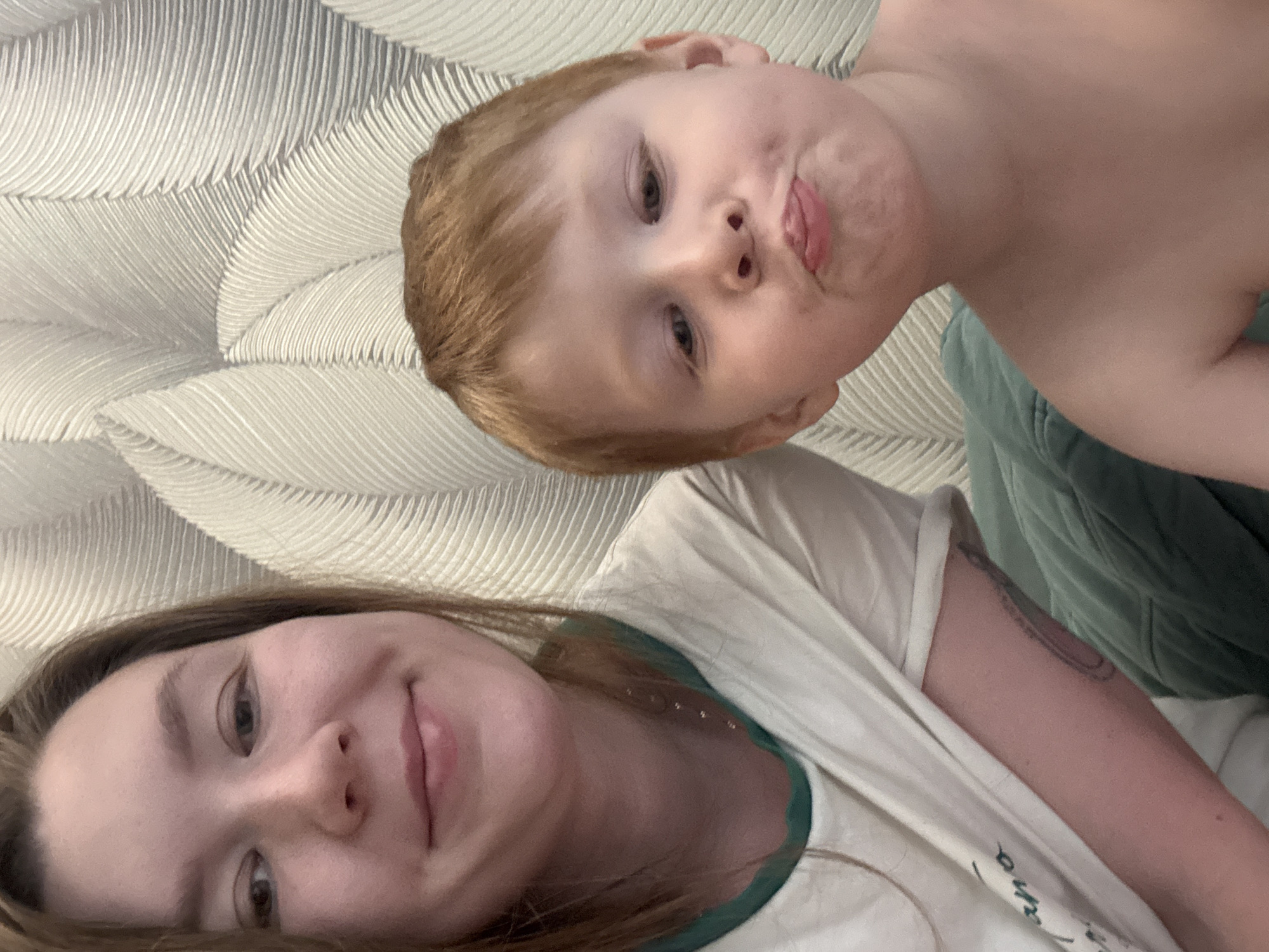 A selfie-style image shows a woman and a young child with red hair. The child is making a playful face with pursed lips and a slightly protruding tongue. The woman is smiling gently. They are indoors against a backdrop of textured, possibly patterned wallpaper. The location cannot be determined as Izhevsk, Russia.
