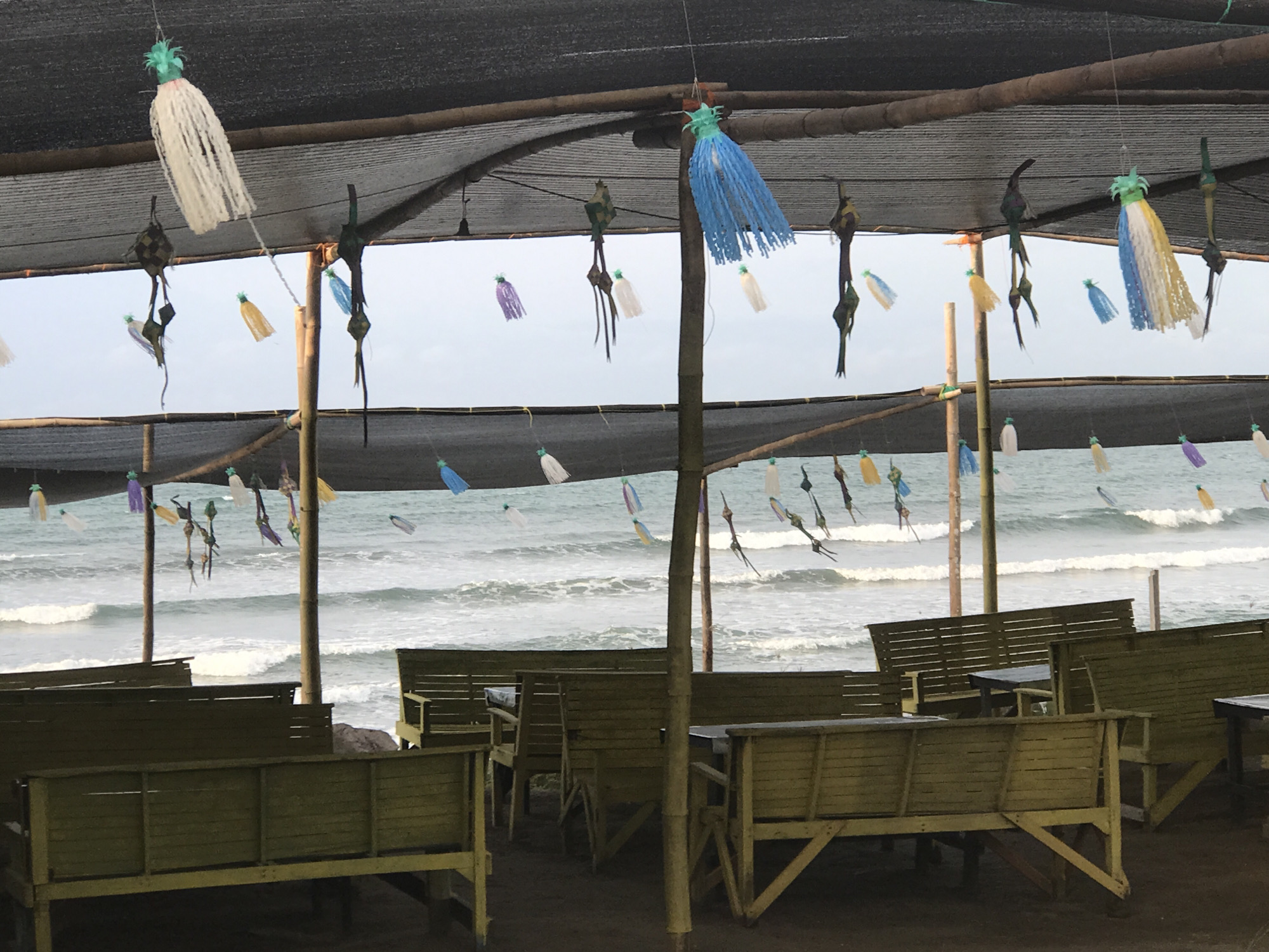 The image shows a covered outdoor seating area with wooden tables and benches facing the ocean. A black mesh netting provides shade overhead. Hanging from the netting are various colorful ornaments, including tassel-like decorations and objects resembling birds. In the background, waves are visible on the ocean, and the sky is overcast. The location is likely a beachside restaurant or cafe. The scene is situated in Lhokseumawe, Indonesia, based on the prompt's indication. There are no people visible in the photograph.