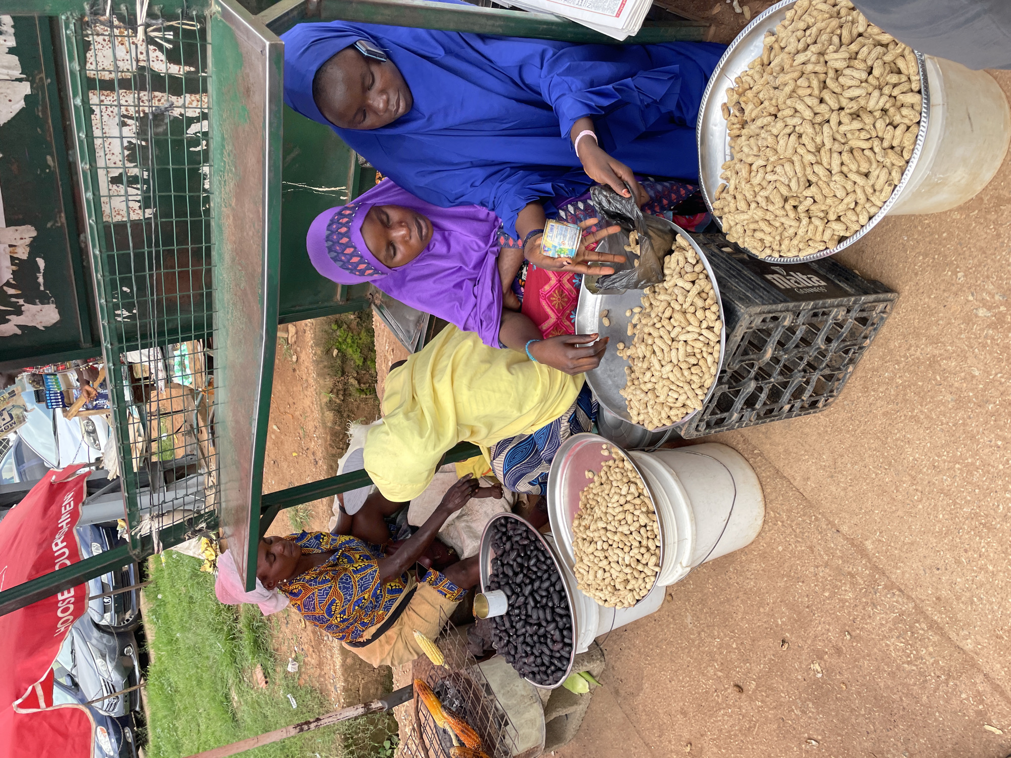 In Bar Jirgi Summa, Nigeria, an outdoor market stall displays a variety of goods. A woman in a vibrant, patterned dress and headscarf sits on the ground, tending to grilled corn on a metal grate. In front of her, large buckets hold piles of food items: a bucket to the left contains dark, round fruits, possibly dates or prunes, while another bucket holds a mound of peanuts in their shells. Two younger women, one in a purple hijab and the other in a blue hijab, are also present at the stall. The woman in the blue hijab appears to be holding a small can and a black plastic bag, possibly preparing to package some of the peanuts for a customer. They are engaged in a transaction, with the woman in purple looking on. The ground is a dusty, reddish-brown earth. In the background, cars are parked on a grassy verge, and a red awning with white lettering, partially obscured, covers part of the stall. A green metal structure, likely part of the market stall's shelving, is visible behind the people.
