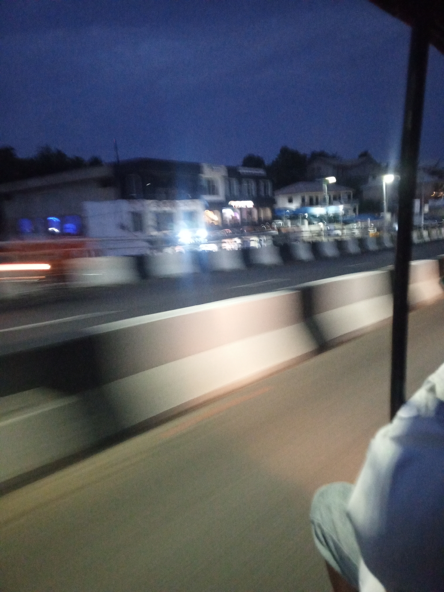 The image depicts an outdoor urban scene at night, possibly taken from a moving vehicle. In the foreground, a person wearing a white shirt and grey pants is visible. The middle ground shows a road with a barrier dividing the lanes. Beyond the barrier, blurred buildings with illuminated storefronts and streetlights suggest a commercial area. Streaks of light indicate moving vehicles. The sky is dark.
The location is the city of Unknown, Unknown.