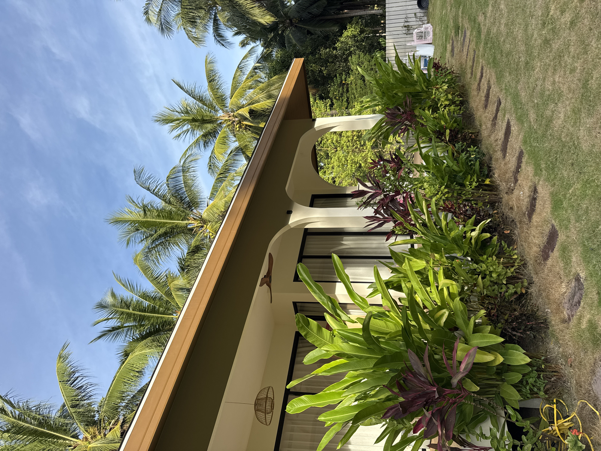 The image depicts the exterior of a building with a brown-trimmed roof, situated in a tropical environment. Lush green foliage, including large-leafed plants and palm trees, surrounds the structure. A pathway of square stepping stones leads across a grassy lawn. The scene is bathed in sunlight, suggesting it is daytime. The architecture includes arched openings and large windows with white curtains. A woven light fixture hangs from the eaves, and a wooden ceiling fan is visible under the overhang. In the background, a white fence and some pink children's play equipment can be seen. The sky above is a clear blue with wispy white clouds. The location is specified as Ko Pha Ngan, Thailand.