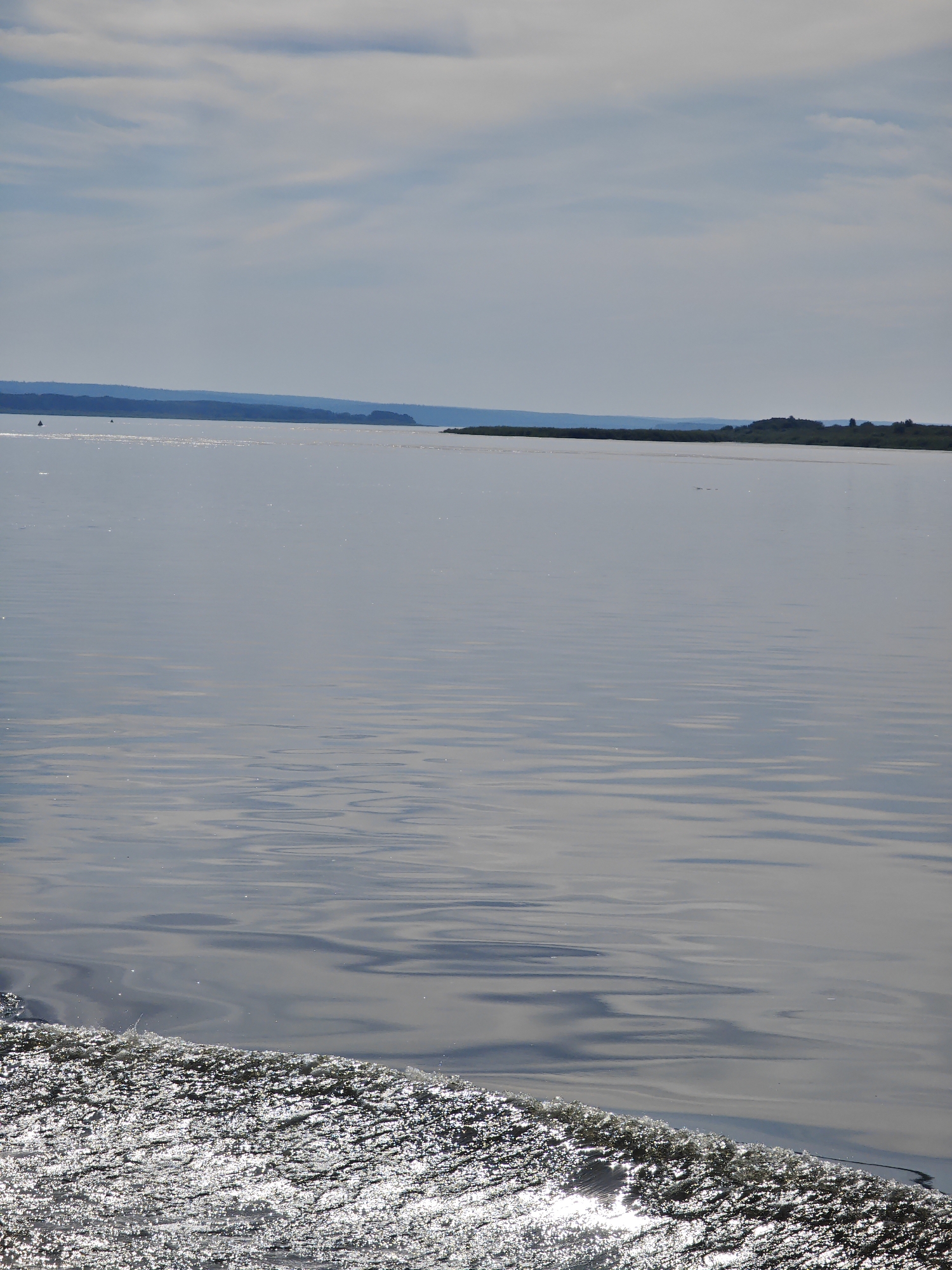 A wide, calm body of water stretches across the frame under a hazy, cloudy sky. The water's surface reflects the light, creating shimmering patterns. In the distance, a dark, forested shoreline with a slight elevation is visible, forming a distinct horizon line. The foreground features a foamy wake, a result of movement through the water, sparkling with sunlight. There are no people or specific objects readily identifiable in the scene, which appears to be a natural landscape. The setting suggests a location along a river or lake in Krivlyak, Russia, characterized by its natural beauty and open water.