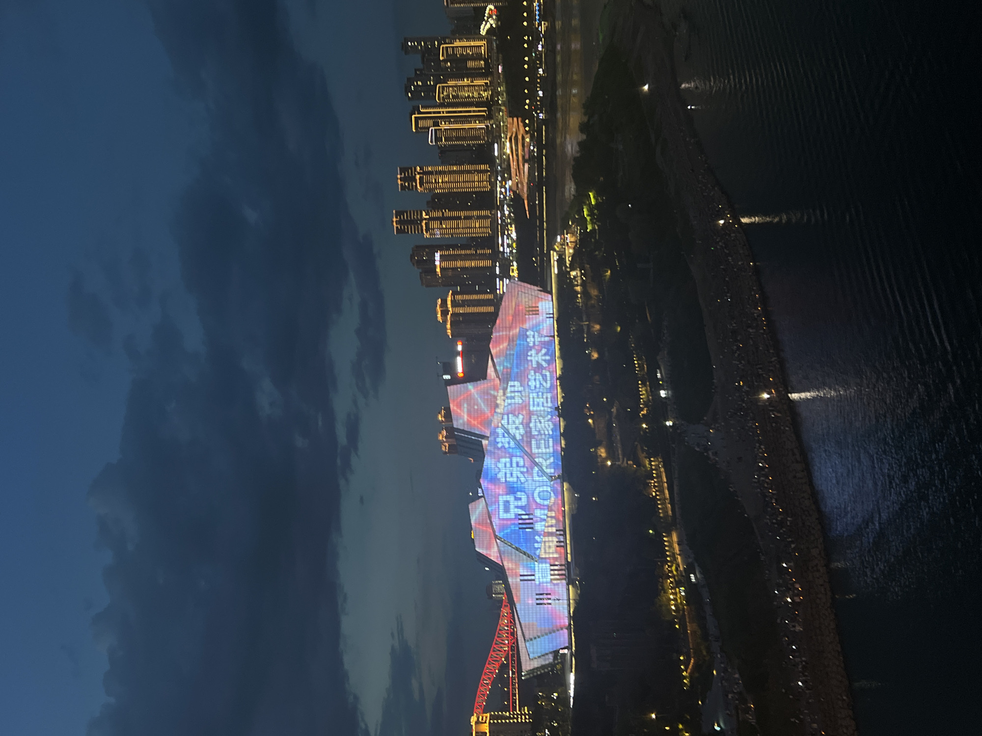 The image shows a nighttime cityscape in Yuzhong District, China, featuring the Yangtze River and illuminated buildings. A large, modern building with a unique architectural design is prominently displayed in the foreground, its facade covered in colorful light projections. Text in Chinese and the English word "MORE" are visible on the building's illuminated surface, suggesting it is a commercial or cultural venue. To the left, a red bridge spans the river, its structure also outlined with lights. In the background, a cluster of tall buildings with illuminated windows forms a dense urban skyline. The river flows between the foreground and the background, reflecting the lights of the city. Along the riverbank in the foreground, a walkway or embankment is visible, lined with numerous small lights and a crowd of people observing the illuminated building. The sky above is dark and cloudy, indicating twilight or nighttime. The overall scene depicts a vibrant and dynamic urban environment at night.