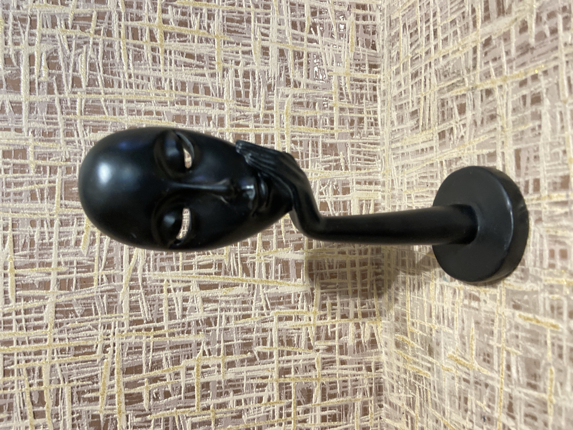 A black, abstract sculpture of a human head supported by a hand is centrally positioned against a textured background. The sculpture, rendered in a matte black finish, features a stylized head with closed or hollow eye sockets, a subtle nose, and a slight, serene smile. A right hand, with defined fingers, cradles the chin and jawline of the head. This head and hand assembly is mounted on a slender, cylindrical black column, which terminates in a flat, circular black base. The background consists of a patterned surface, likely wallpaper, characterized by a dense, interwoven or grid-like arrangement of raised light beige and yellowish-gold lines over a muted reddish-brown base. The pattern is consistent across the vertical and horizontal planes of the background. There are no individuals, actions, or interactions depicted. The lighting is even, illuminating the textured background and the smooth surface of the sculpture. The image contains no discernible visual elements to determine its geographic location as Tuymazy, Russia, or any other specific city.