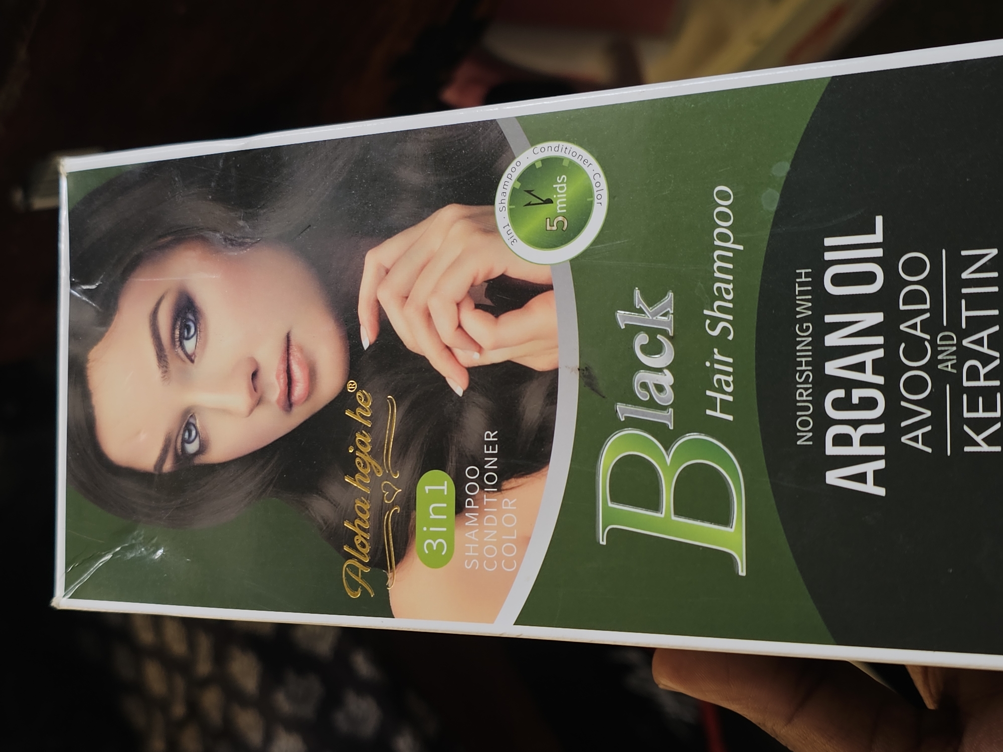 The image is a close-up, eye-level photograph of the front of a product box for "Aloha heja he" Black Hair Shampoo. The box features a woman's face and hair, along with product details such as "3in1 SHAMPOO CONDITIONER COLOR" and "5 mids." The lower portion of the box highlights "NOURISHING WITH ARGAN OIL, AVOCADO AND KERATIN." The background of the image is dark and out of focus, suggesting the product is being held or displayed in a dimly lit indoor environment. There is no discernible indication of the location being the city of Dar es Salaam, Tanzania, or any specific city in general.