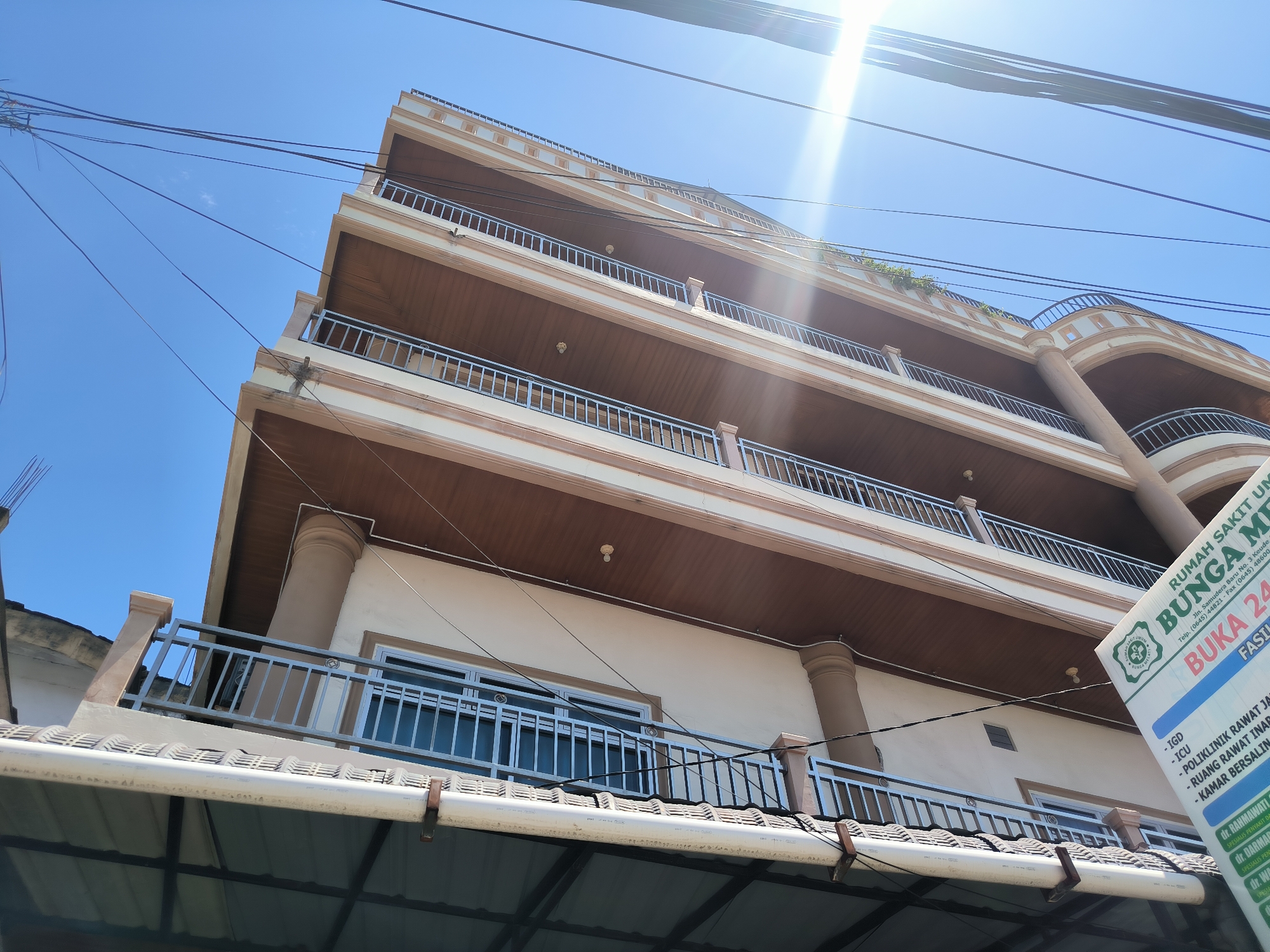 The image is a low-angle shot of a multi-story building in Lhokseumawe, Indonesia. The building has a tan exterior with brown balconies on each floor. A large sign is partially visible on the right side of the frame with the words "Rumah Sakit Umum Bunga Melati" (General Hospital Flower Jasmine) and the words "Buka 24" (Open 24). Overhead power lines cross the sky. The sky is bright blue, and the sun is visible in the upper right corner, creating a lens flare.