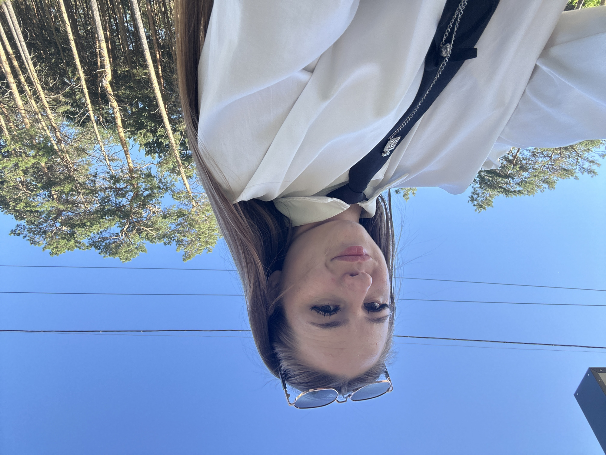 An outdoor, waist-up, eye-level, full shot captures a young woman from the chest up, posing for a selfie. She is wearing a white button-down shirt with a black tie that has a silver charm attached. Her sunglasses are perched on her head, and she is looking directly at the camera with a slight smile. The background consists of a clear blue sky with power lines strung across it, and a stand of pine trees to the right. The scene is bathed in bright sunlight. The location is unknown.