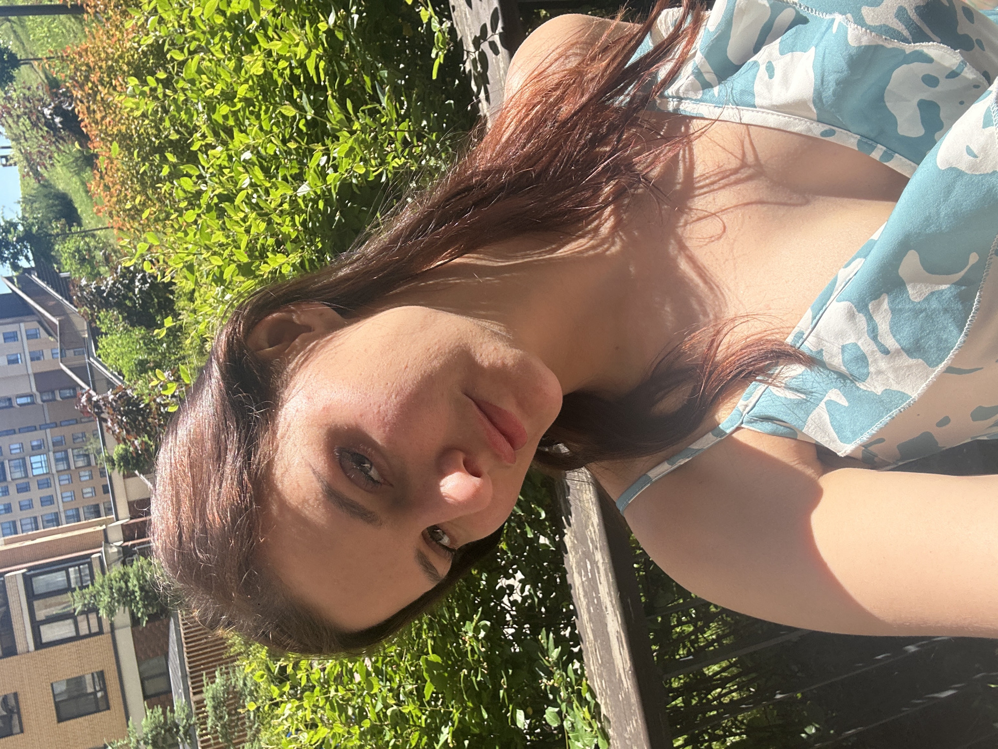 In a medium shot, a young woman with long, dark brown hair is photographed outdoors on a sunny day, looking towards the camera. She is wearing a light blue and white patterned dress with ruffled straps. Her head is tilted slightly to her left, and her gaze is directed forward. The background features lush green foliage, suggesting a natural or park-like setting. Behind the greenery, a multi-story building with an angular architectural style is visible, and a paved pathway with steps ascends a grassy slope on the right side of the frame. A wooden railing or bench is positioned in the foreground, partially obscuring the lower part of the image. The sunlight casts shadows and highlights on the woman's face and arms, indicating a bright, clear day.