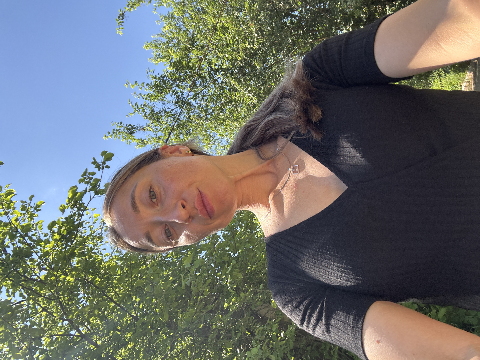 A woman is taking a selfie outdoors. She is wearing a black, short-sleeved, v-neck top and a silver pendant necklace. She has her hair pulled back in a ponytail and is wearing small gold earrings. The background is out of focus and consists of green trees and a blue sky. The image was taken during the day. The location is Chaikovsky, Russia.