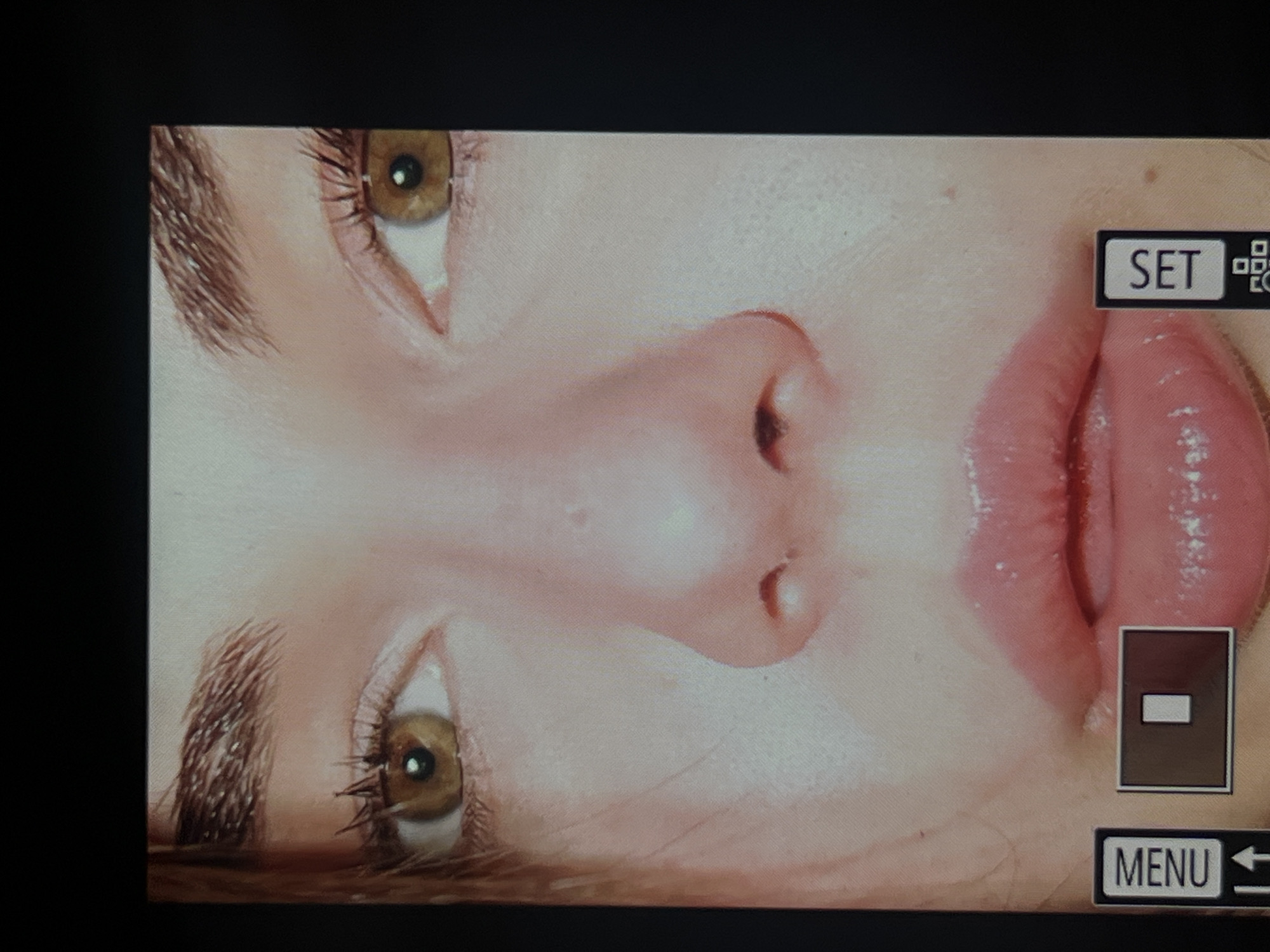 The image shows a close-up of a woman's face, rotated approximately 90 degrees counterclockwise. The visible features include two eyes with brown irises, eyebrows, a nose, and lips. The lips appear to have gloss or a similar product applied. Overlaid on the image are camera interface elements: "SET" with a QR code icon, a rectangular icon with a horizontal line inside, and "MENU" with a left-pointing arrow. There is no information in the image indicating that it was taken in Saint Petersburg, Russia.