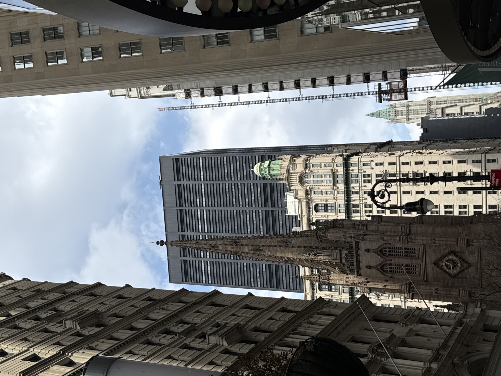 The image depicts a street view in New York City, USA, taken from a low angle looking upwards. The composition is dominated by the architectural structures of several buildings, which stretch towards a sky with scattered clouds. On the lower left and right, building facades are visible with rows of windows, indicating dense urban construction. Prominently featured is the spire of Trinity Church, an iconic landmark in the Financial District, situated near a modern, glass-paneled skyscraper. In the upper portion of the frame, there is construction scaffolding against the backdrop of another building, suggesting ongoing urban development or maintenance. Street lighting fixtures are also visible, adding to the urban environment.