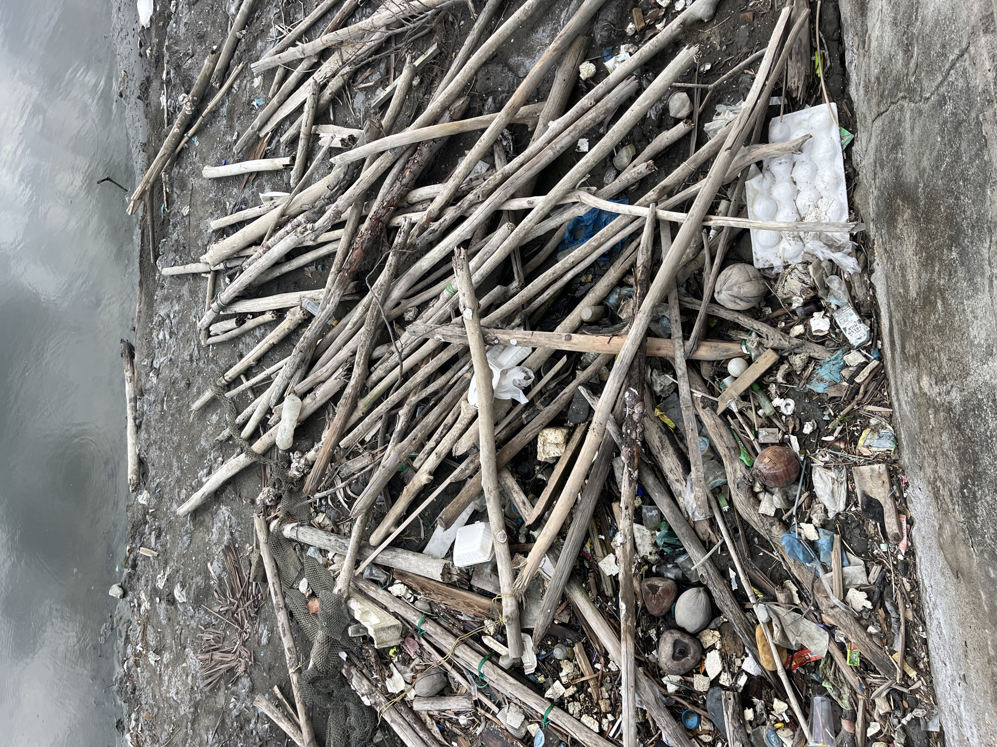 The image depicts a polluted shoreline in Lhokseumawe, Indonesia. The foreground is dominated by a chaotic pile of driftwood and trash. The trash includes plastic bottles, plastic bags, Styrofoam pieces, and discarded food containers. Some coconuts are visible amongst the debris. To the left of the image is a body of water, and to the right, there is a concrete wall. There are no people visible in the scene.