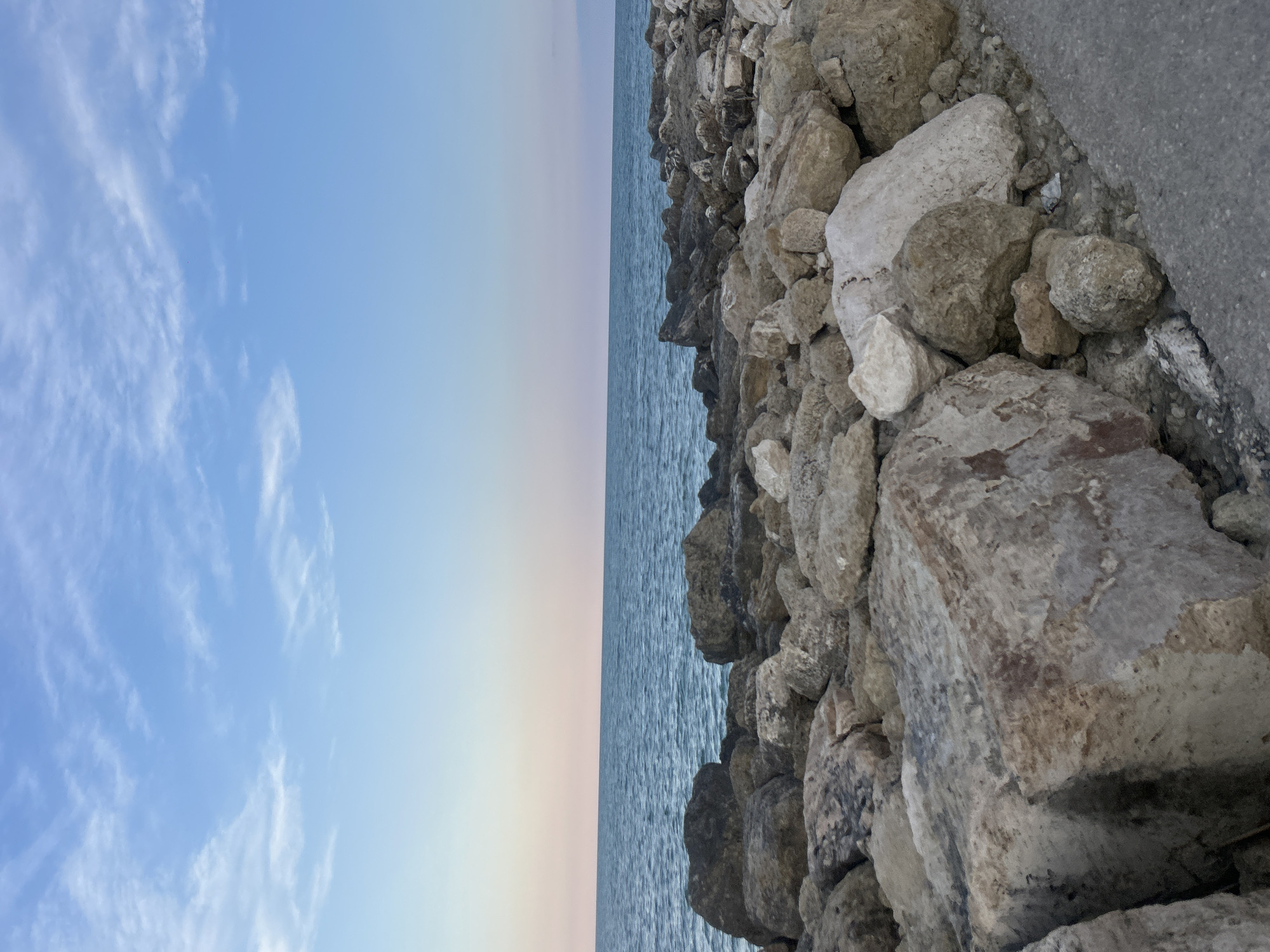 The image shows a rocky coastline with the sea on the right side of the frame. The sky is a pale blue with wispy white clouds. The water is a calm, deep blue. The rocks in the foreground are various sizes and colors, ranging from light gray to brown and white. There appears to be a paved path or sidewalk on the far right of the image. This scene is consistent with the rocky breakwaters found along the coast of Grottammare, Italy. There are no people or specific actions depicted in the image.