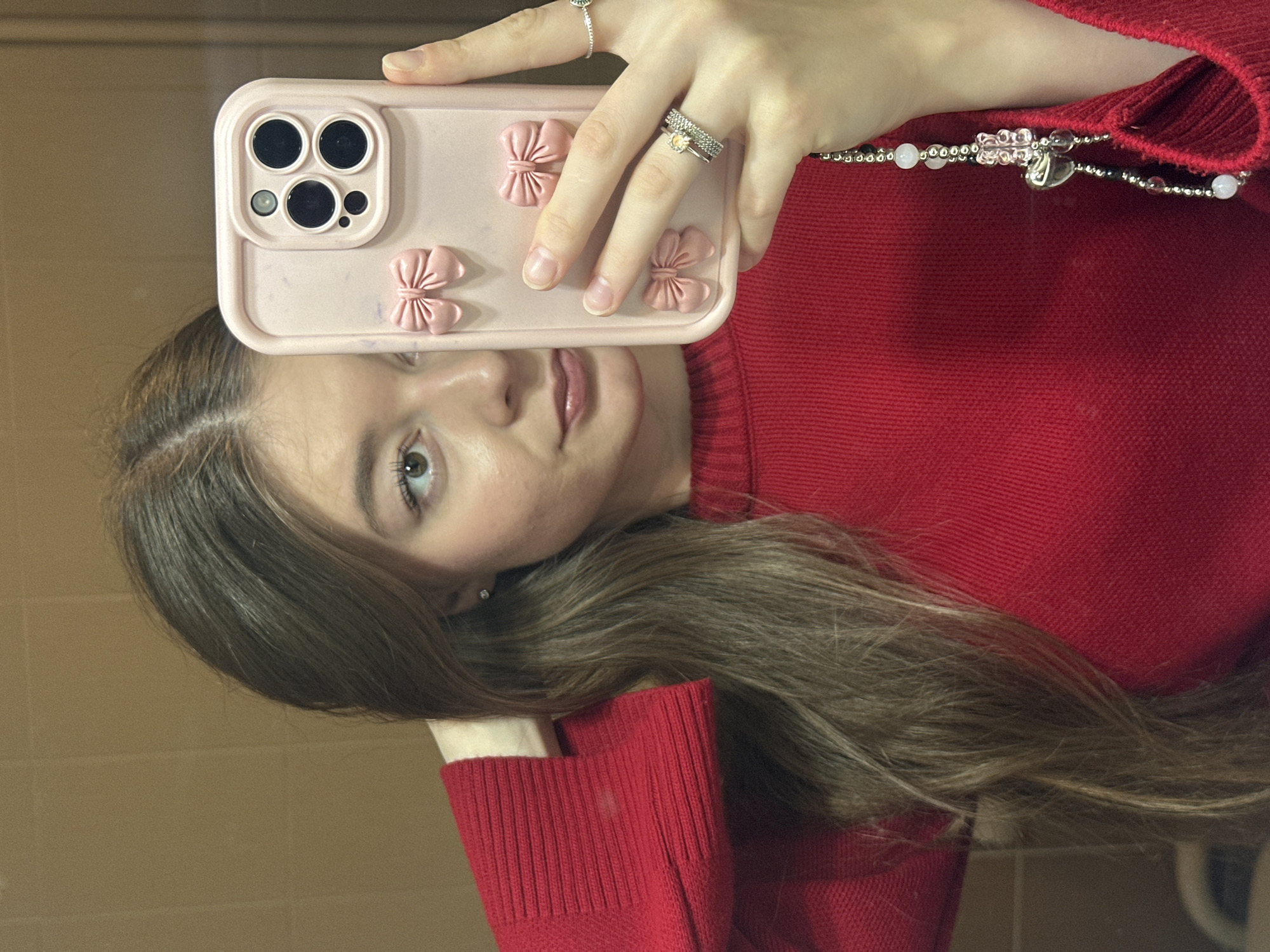 A young woman is taking a mirror selfie indoors, holding a light pink smartphone with a decorative case featuring three prominent bow-shaped embellishments. Her face is visible in the reflection, showing a neutral expression as she looks slightly upwards towards the camera lens. She has long, straight brown hair, parted subtly, and light-colored eyes. She is wearing a red ribbed sweater, and a multi-strand beaded necklace or bracelet with silver beads, clear/white beads, and a heart-shaped charm is visible around her neck/collarbone area.
On her left hand, which is holding the phone, several rings are visible: a thin silver band with a small, light-colored stone on her ring finger, an adjacent silver band, and a delicate silver ring with a green stone on her index finger. Her fingernails are natural and unpainted. The phone itself appears to be a model with a triple camera system and a flash, typical of recent iPhone models. The background reflected in the mirror suggests a plain, light brown or tan wall, possibly tiled. The lighting appears to be indoor, and there are no discernible specific location cues or visible text within the image.
