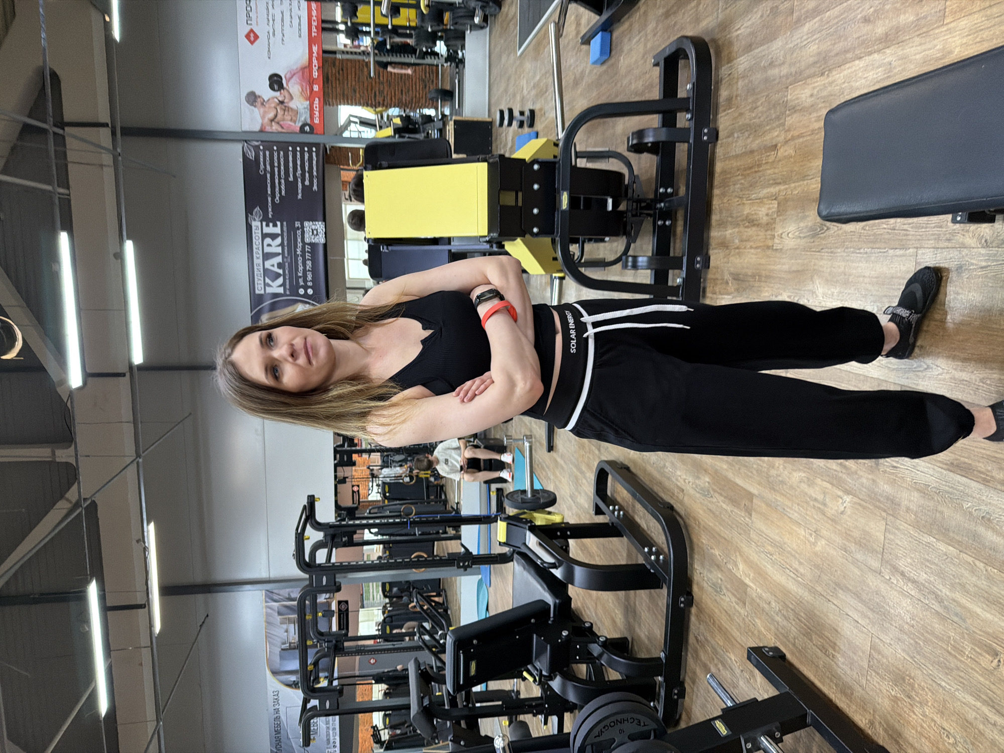 The image shows a woman standing in a gym in Chaikovsky, Russia. She is wearing a black top and black sweatpants with white stripes. She has her arms crossed. The gym has various workout machines and equipment. There is a sign for a beauty studio called "KARE" located on Karl Marx street, number 31. The phone number for the studio is 89617587777. Another sign promotes fitness training.