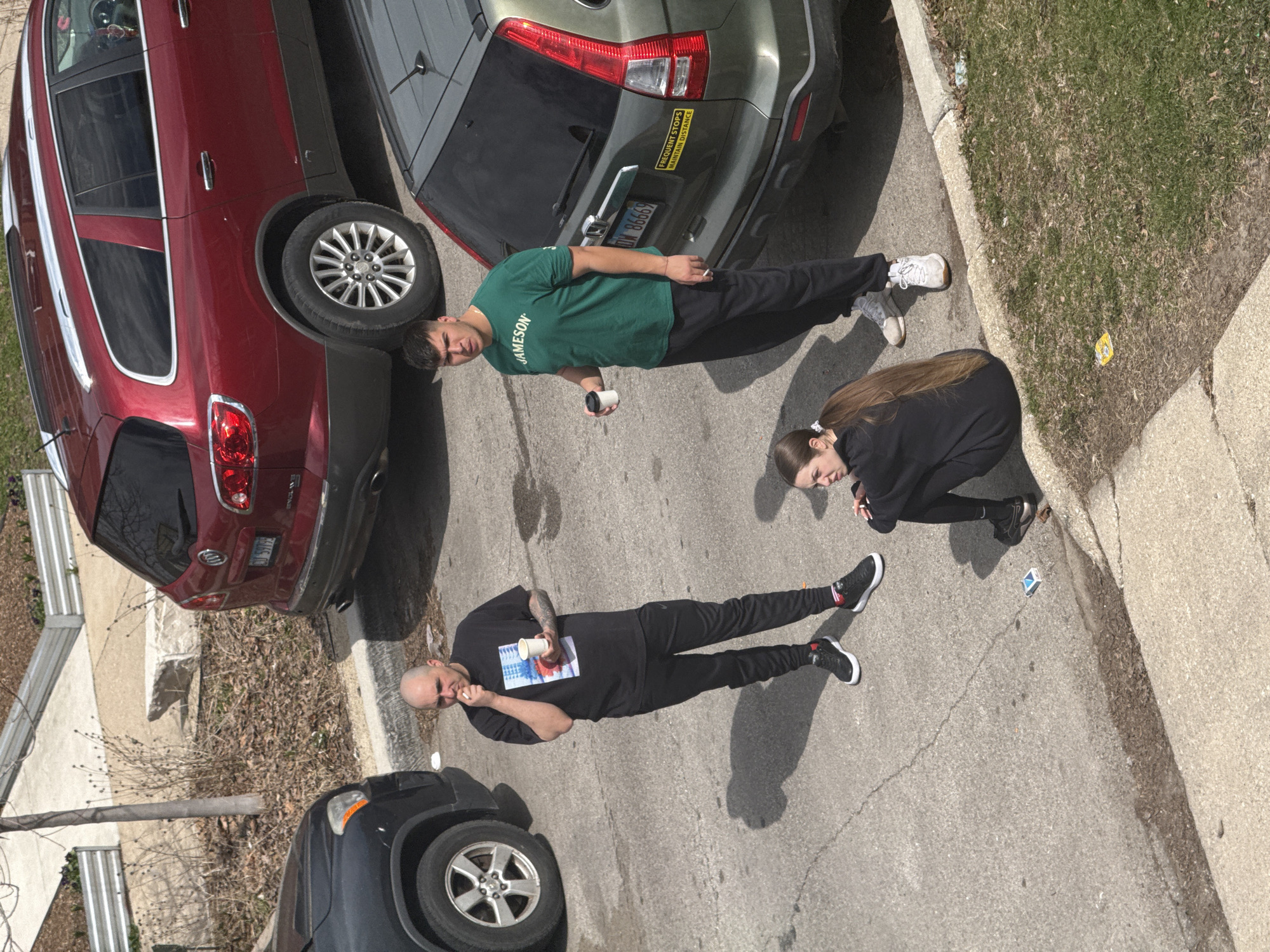 The image shows a street scene in an urban area, likely Chicago, United States of America, based on car models and style. Three people are present. A man wearing a green shirt with "Jameson" on it is standing near a green SUV. A woman wearing a black outfit is crouching near the sidewalk. Another man wearing a black shirt and pants is standing near a black SUV. Both men are holding white cups. There are several parked cars, including a red SUV. The environment includes a street, a sidewalk, and grass along the edge. The weather is sunny. The sidewalk has a small, blue trash item.