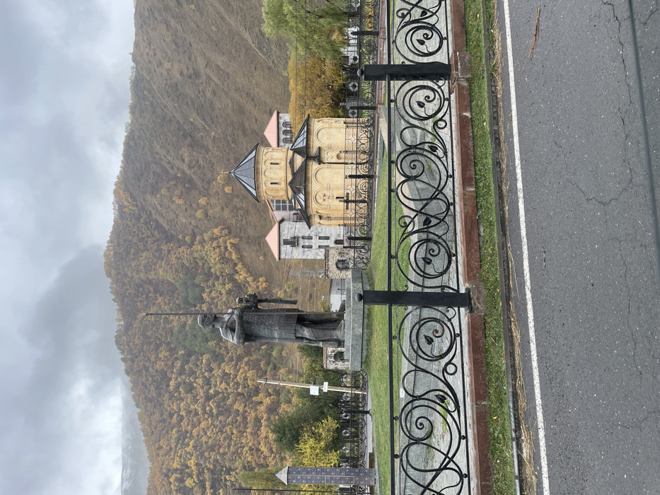 The image depicts a rural mountainous scene in Sno, Georgia, under an overcast sky. In the immediate foreground, a dark asphalt road with a solid white line to the right is visible. Directly behind the road is a low, reddish-brown curb topped with a black wrought iron fence featuring intricate scrollwork and leaf motifs.
Behind the fence lies a manicured grassy area. Centered within this area is a large, dark-colored statue of a standing male figure, dressed in armor, holding a long spear-like object vertically in his right hand and a smaller object (possibly a scroll or horn) in his left hand. The statue stands on a multi-tiered, light-grey stone pedestal. To the right of the statue, a light yellow-beige stone church with a central dark-domed roof and two smaller side domes is prominent. The church's facade features ornate crosses and is surrounded by its own dark, decorative metal fence. Behind the church, a multi-story building with light walls and distinctive reddish-orange pitched roofs is partially visible. To the left of the statue, another tall, narrow, dark monument with tiled patterns is partially visible.
The background is dominated by steep mountainsides covered extensively with deciduous trees displaying vibrant yellow and orange autumn foliage. Patches of darker green trees are also interspersed. The upper slopes of the mountains are obscured by low-hanging clouds or mist. The sky above is entirely overcast and grey. No individuals are visible in the scene.