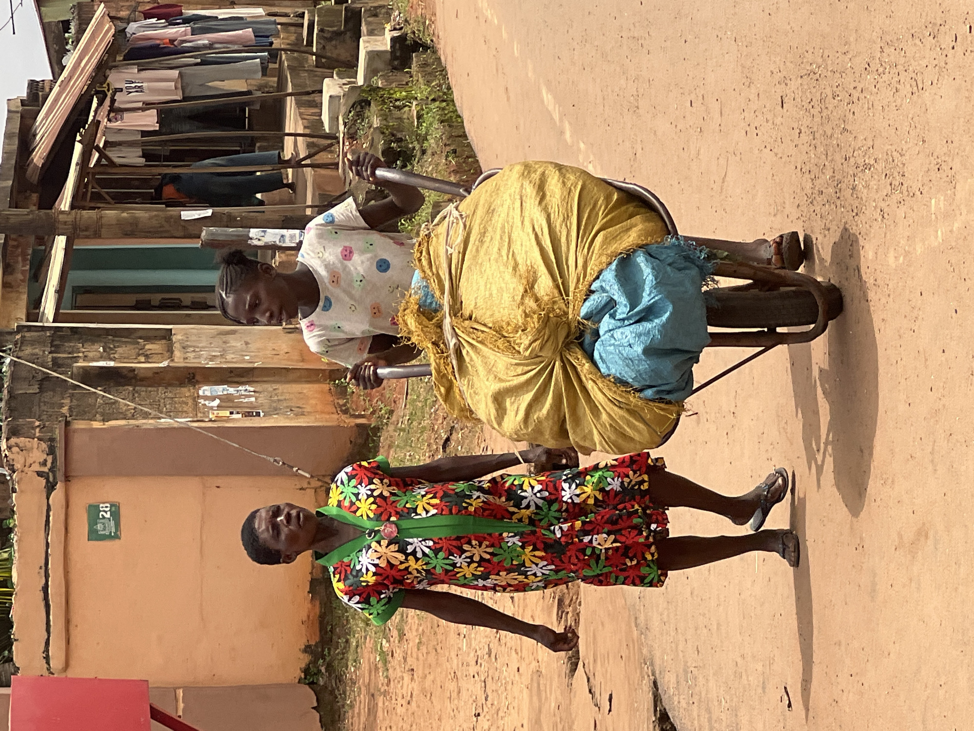 In Nkpor, Nigeria, a young woman pushes a wheelbarrow loaded with a large yellow bag and a blue bundle down a dusty, reddish-brown street. She wears a white t-shirt with a pattern of colorful smiley faces. To her left, an older woman walks beside her, wearing a vibrant floral dress in red, yellow, green, and white with a green collar and trim. The background shows a street scene with buildings, one painted a peachy-orange with a green sign displaying the number "28," and another with a corrugated metal roof and clothes hanging on a line. The overall environment is an outdoor, urban setting.