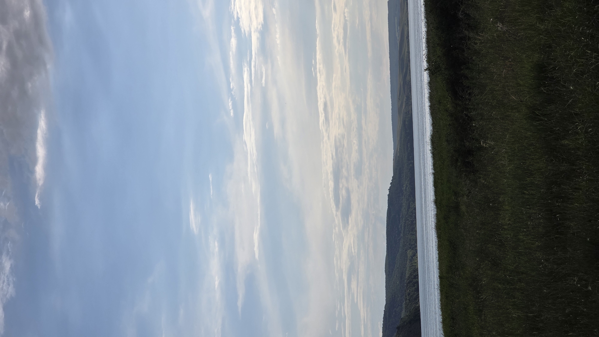 This is a photograph of a natural landscape. The scene is dominated by a vast expanse of blue sky with scattered, wispy white clouds. To the right of the sky, a body of water stretches across the frame, reflecting the sky and appearing as a bright, narrow band. Beyond the water, a densely vegetated hillside rises, covered in trees and green foliage. The foreground is a field of tall, dark green grass, occupying the right side of the image. The perspective is from a slightly elevated position looking out over the scene. There are no people or man-made objects visible. The lighting suggests it is daytime, likely late afternoon or early evening given the soft quality of the light. The location is identified as Kupoyarovo, Russia, hinting at a rural or natural setting. The overall mood is serene and peaceful, emphasizing the beauty of the natural environment. There is no visible text in the image.