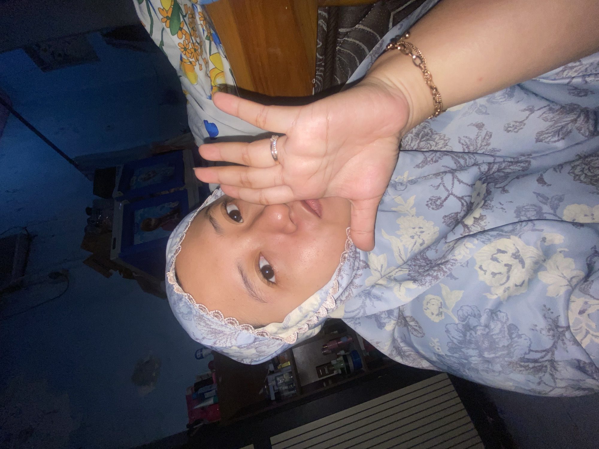 A woman wearing a light blue floral hijab and matching dress is seen from the chest up, holding her left hand up to the camera, obscuring part of her face. She is wearing a silver ring on her left ring finger and a gold bracelet on her left wrist. Her expression is difficult to discern due to her hand placement, but her eyes are open and looking at the camera. The background is dimly lit and appears to be an indoor setting. To the left, shelves with various items are visible, including bottles and framed pictures. To the right, bedding with a floral pattern is partially visible, and a wooden surface suggests furniture. The overall ambiance is casual and personal. The image was taken in Gelam, Indonesia.