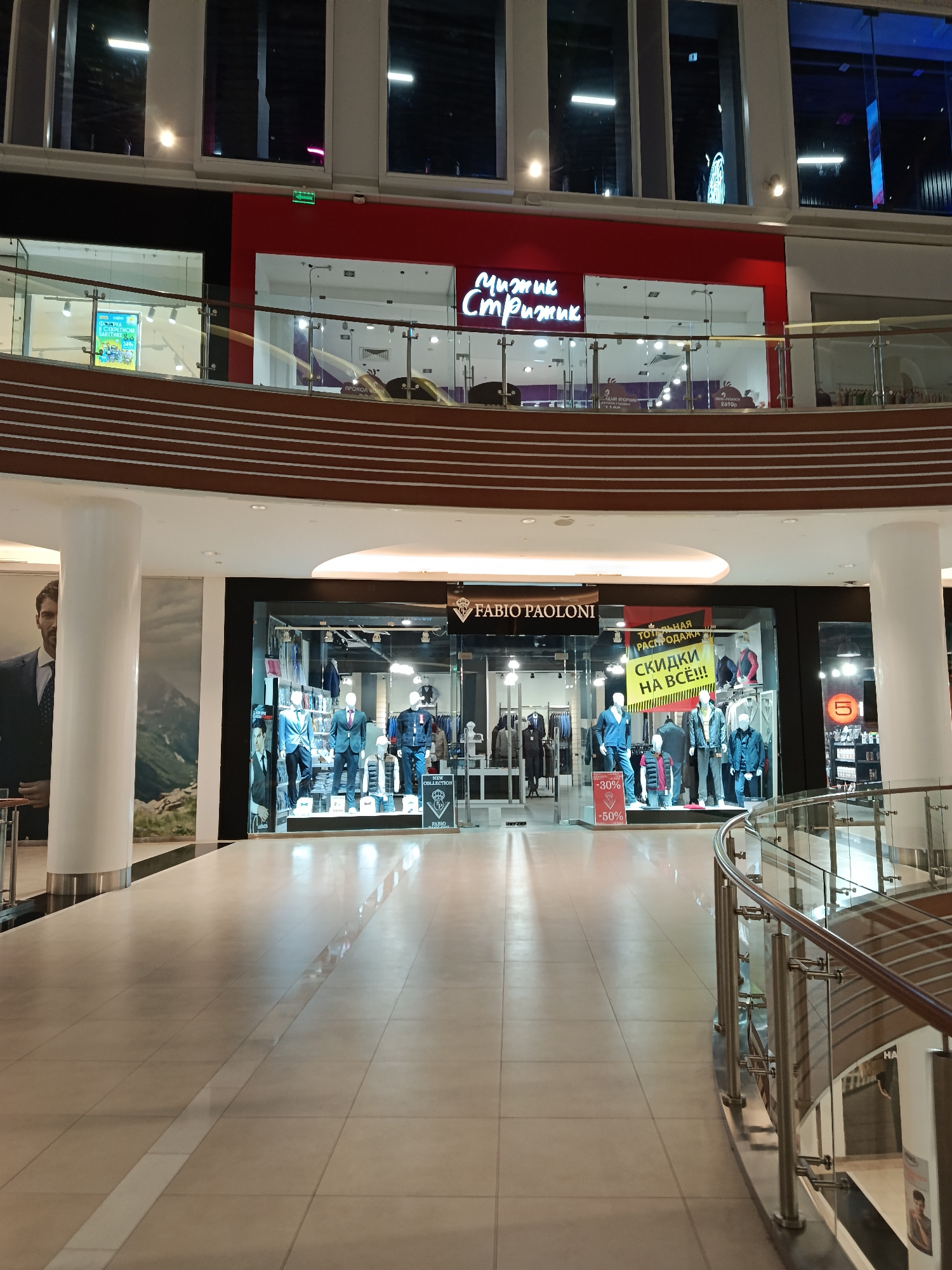 This is an indoor shot of a shopping mall, likely in the city of Unknown, Unknown. The view is a corridor or walkway, with tiled flooring, leading towards a store named "FABIO PAOLONI". This store appears to be selling men's clothing, with mannequins dressed in suits and casual wear visible through the glass storefront. Sales signs indicating discounts, such as "-30%" and "-50%", are prominently displayed.
Above "FABIO PAOLONI", on an upper level, there is another store with a red facade and a sign in Cyrillic script, possibly "Чижик Стрижик".
The scene is well-lit with recessed ceiling lights. A modern glass and metal railing lines the right side of the corridor, adding a sense of spaciousness and depth. A large advertisement featuring a man in a suit is visible on the left, partially obscured by a column.