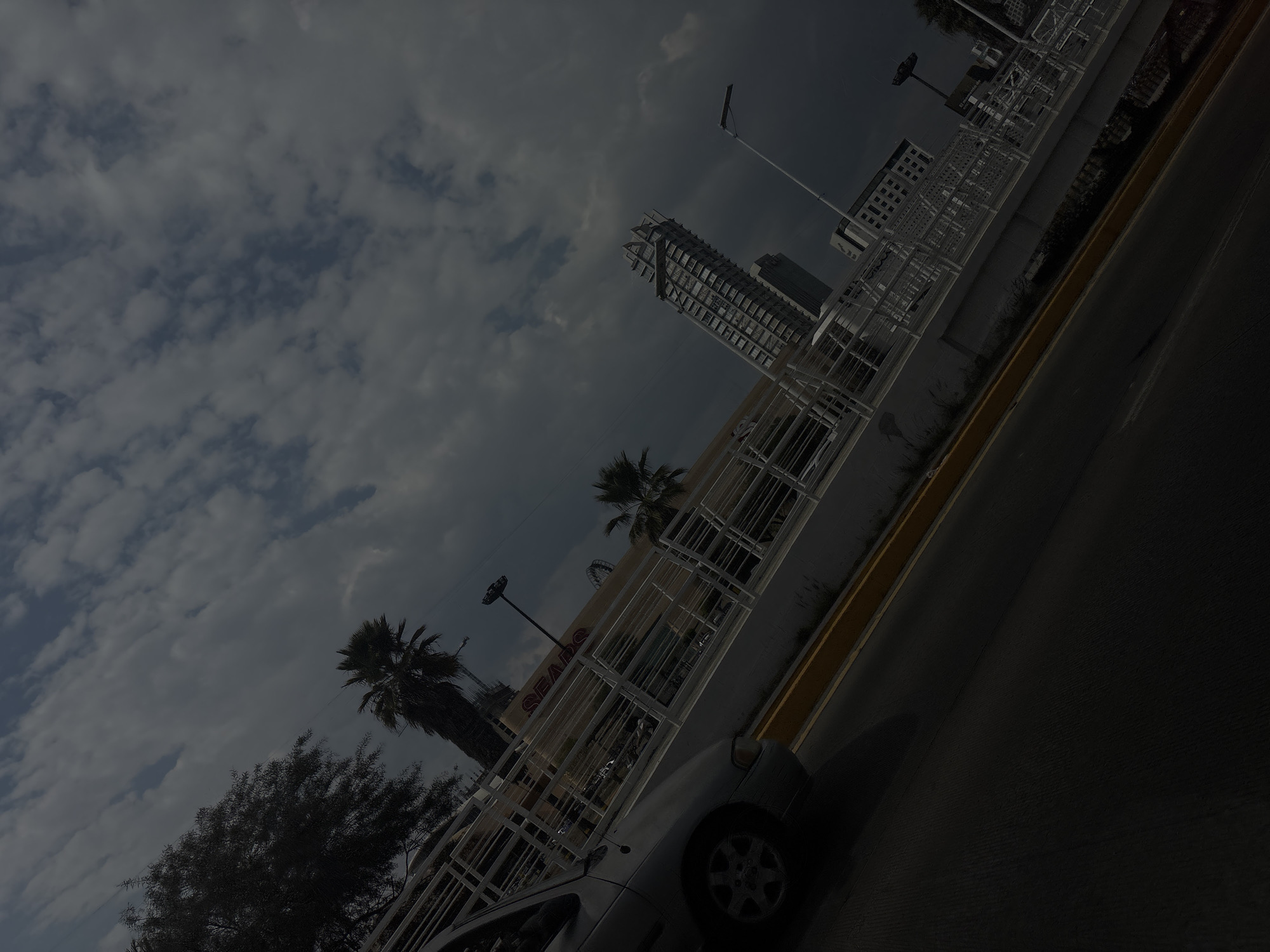 The image, captured from a low angle, depicts an urban scene under a cloudy sky. The foreground features a silver-colored vehicle, partially visible, parked alongside a street. The street has a yellow curb. In the background, palm trees and deciduous trees are visible, along with a building facade displaying the "SEARS" logo. Further back, modern architecture, including a high-rise building under construction, can be seen. The scene is likely in Puebla, Mexico.