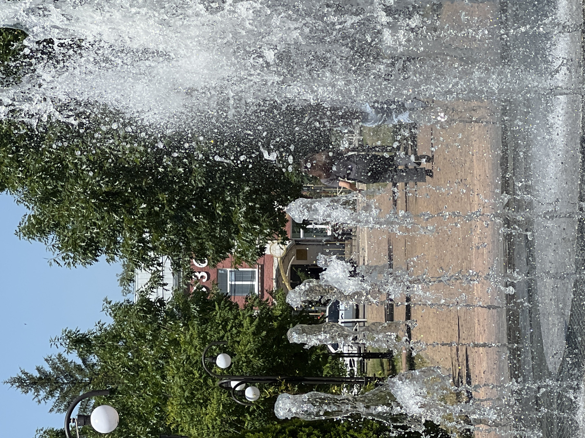 In the foreground, water from a fountain sprays across the frame, obscuring the background. Behind the water, a woman with long, dark hair walks away from the viewer on a paved pathway. She is wearing a dark t-shirt and dark pants. To the left, lampposts with white spherical lights are visible. Further in the background, a red brick building with the number "36" in white lettering is visible, surrounded by green trees. Cars are parked along the street in front of the building. The scene appears to be in an outdoor public space.