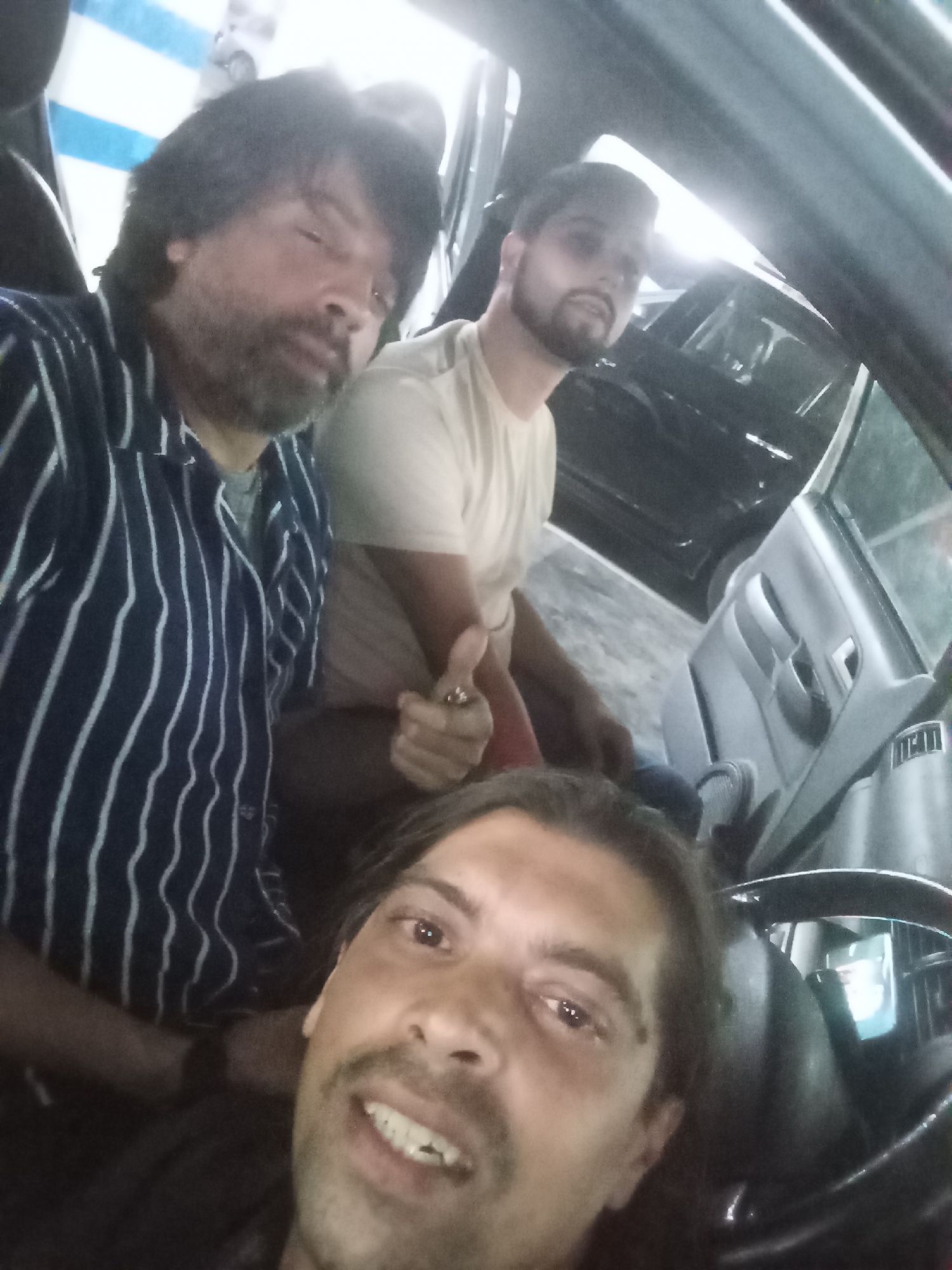 The image depicts three men inside a car, possibly in Buenos Aires, Argentina. The car appears to be parked, potentially in an urban or residential area. The man in the foreground is taking a selfie, capturing himself and the two men behind him. The man in the middle is giving a thumbs up. The car's interior is visible, including the dashboard, steering wheel, and door panel. Another car is seen outside the car they are in. The lighting indicates it could be daytime or a well-lit indoor space.