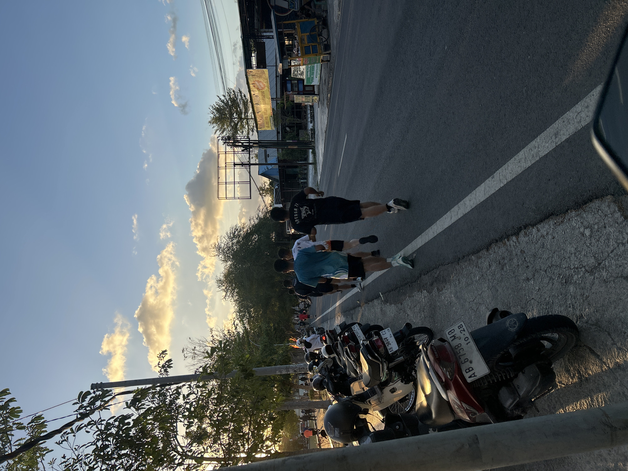 In Wonosari, Indonesia, at approximately 5:30 PM, four men are running along the right side of a paved road. The sun is setting, casting a warm glow on the scene and creating long shadows. The sky is clear with a few scattered clouds. On the left side of the road, several motorcycles are parked along the sidewalk. Trees line the road, and in the background, there are buildings and shops. One of the runners is wearing a blue t-shirt, another a black t-shirt with white text, and the other two are wearing white t-shirts. They are all running in the same direction. The environment appears to be a street in a city or town.