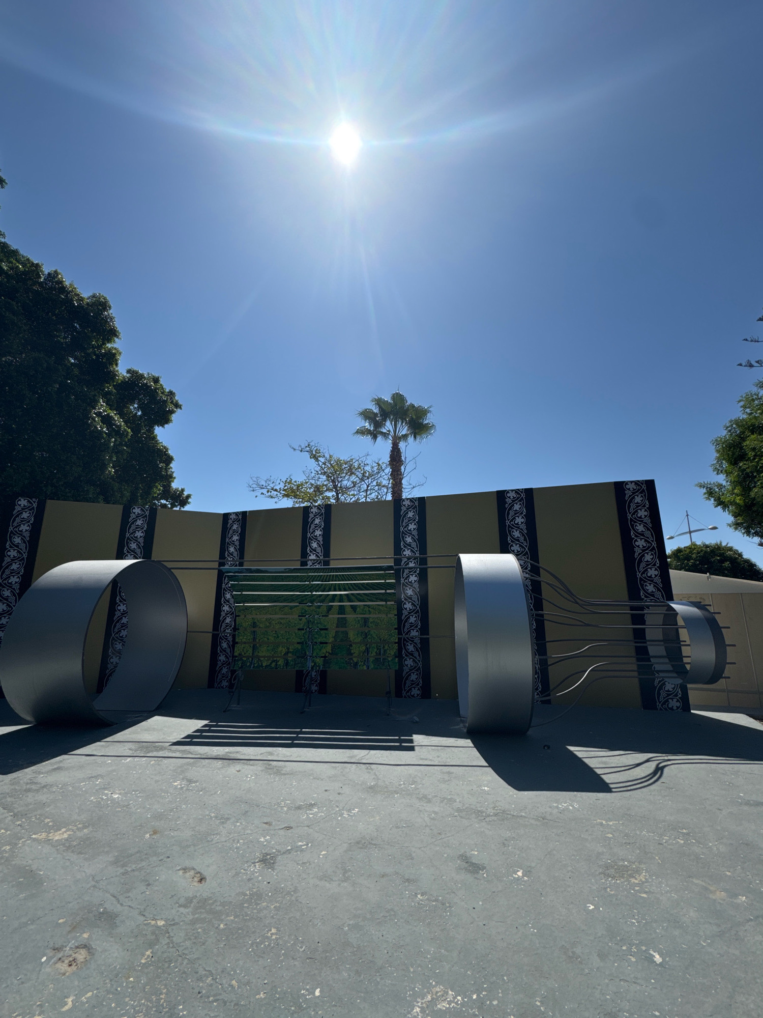 A wide-angle, eye-level, outdoor shot captures a large, modern art installation under a bright, sunny sky. The installation is centered, featuring two large, silver, cylindrical structures at either end. These cylinders are connected by a series of thin, curved metal rods. Behind the installation is a yellowish wall with decorative black and white patterns running vertically. In the middle section of the installation, a green mural depicting a vineyard scene is visible. Trees, including a palm tree, and a street light are seen in the background. The ground in the foreground is a concrete surface with visible wear and tear. The sun is high in the sky, casting strong shadows from the art installation onto the concrete. The overall environment appears to be an urban outdoor space. The location of this art installation is confirmed to be in Limassol, Cyprus.