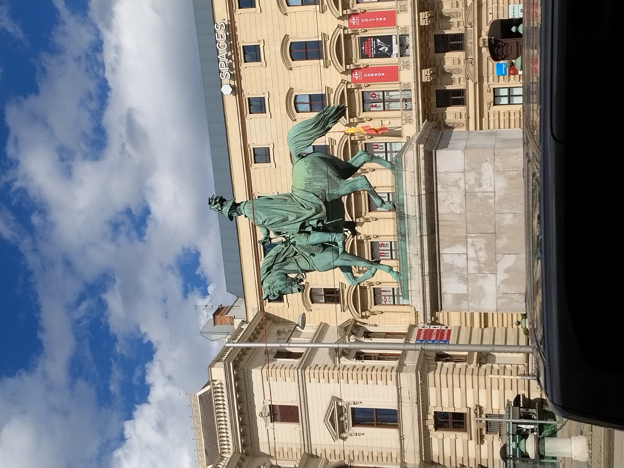 This is a full-frame, eye-level, outdoor, slightly low-angle, bright, daytime, vertical shot of a bronze equestrian statue in Vienna, Austria. The statue depicts a man in military attire with a flowing cape riding a horse. The statue is on a light-colored stone pedestal. Behind the statue is a light-yellow building with multiple windows, some of which are arched. Above the building, the sky is blue with scattered white clouds. A portion of a dark vehicle is visible in the foreground. There are several red banners hanging from the building with text and images on them. A sign with text, including "SPACES," is visible on the top of the building. Other signs indicating directions are also visible on a lamppost near the statue.