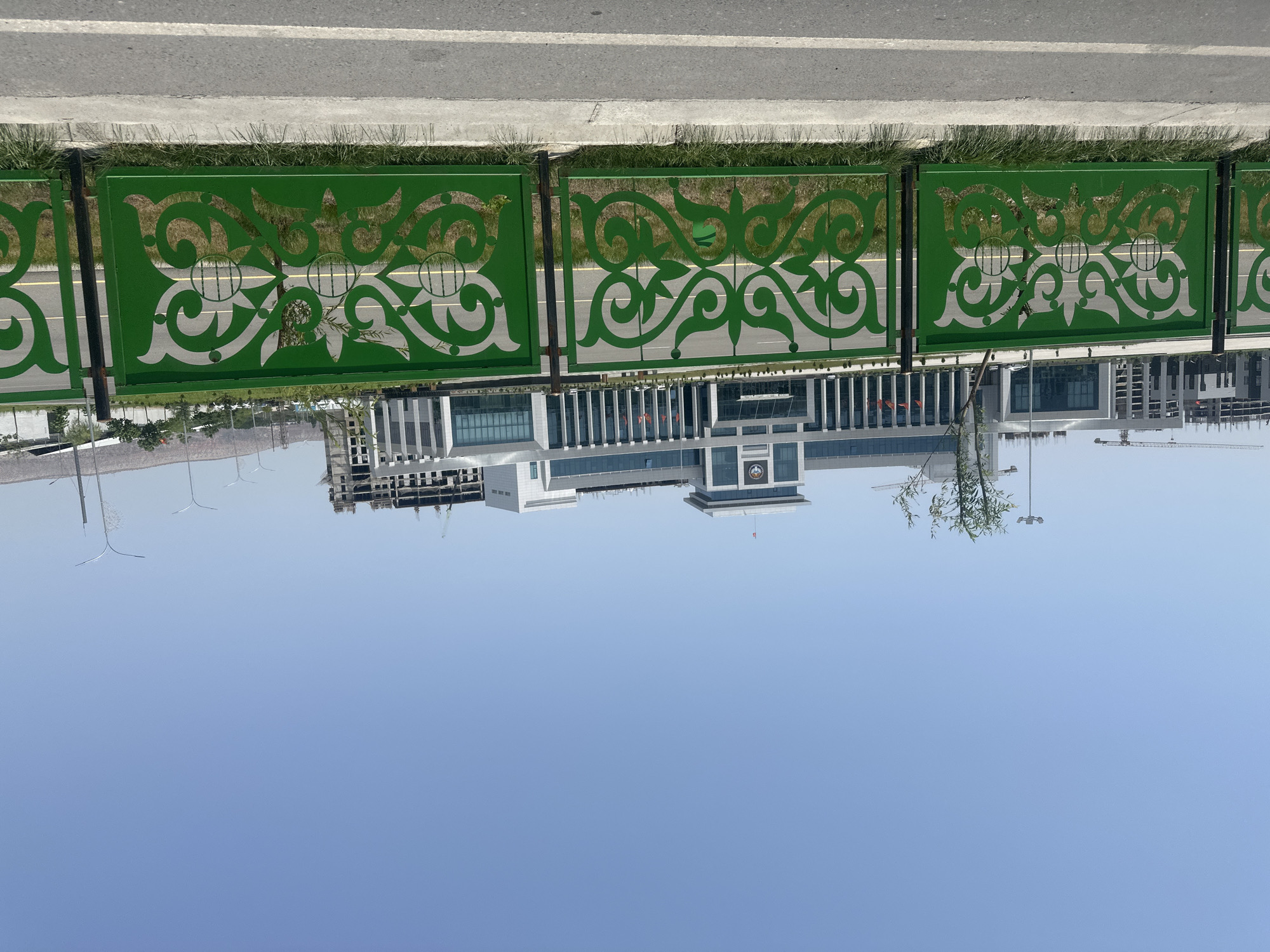 The image depicts a modern building complex behind a decorative green fence with ornate patterns. The fence is metallic and features intricate swirling designs and circular motifs, with glimpses of green grass and some vegetation visible through its openings. In the background, the building is multi-story with a predominantly glass facade and a central section topped with a square structure. There are flags visible on the building. To the left and right of the main building, other structures are present, some appearing to be under construction with visible cranes. The sky is a clear, light blue. The foreground shows a paved road with a white line marking. The scene is set outdoors under daylight. The location is identified as the city of Manas, Kyrgyzstan.