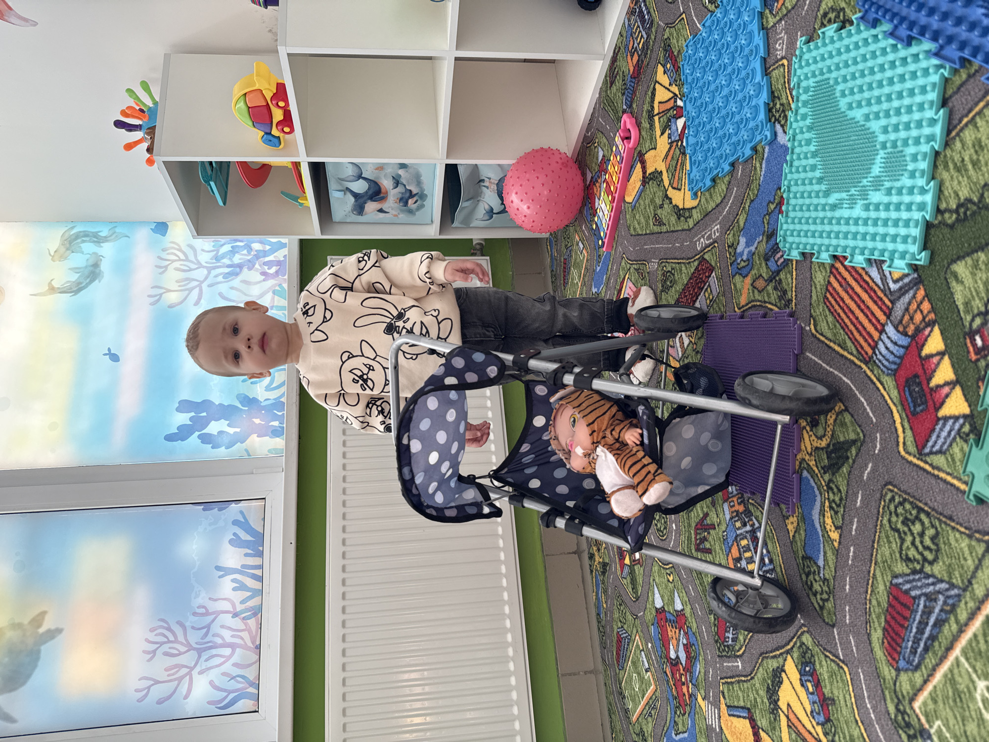 The image shows a toddler standing next to a doll stroller with a tiger doll inside. The toddler is wearing a patterned sweater and dark pants. The stroller is positioned on a colorful carpet with a road pattern. Behind the child, there is a white shelving unit with various toys. To the left, a window with ocean-themed decals can be seen. A white radiator is positioned between the window and the carpet. The location cannot be confirmed as Rusanovo, Russia, from the provided image.